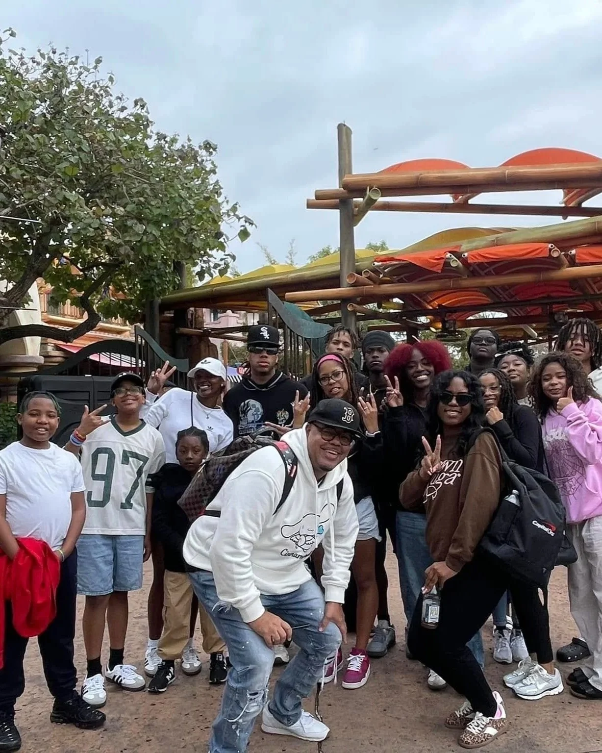 Group of people posing for a photo at an amusement park or outdoor venue, with some making peace signs or other gestures, and colorful rides or structures in the background.