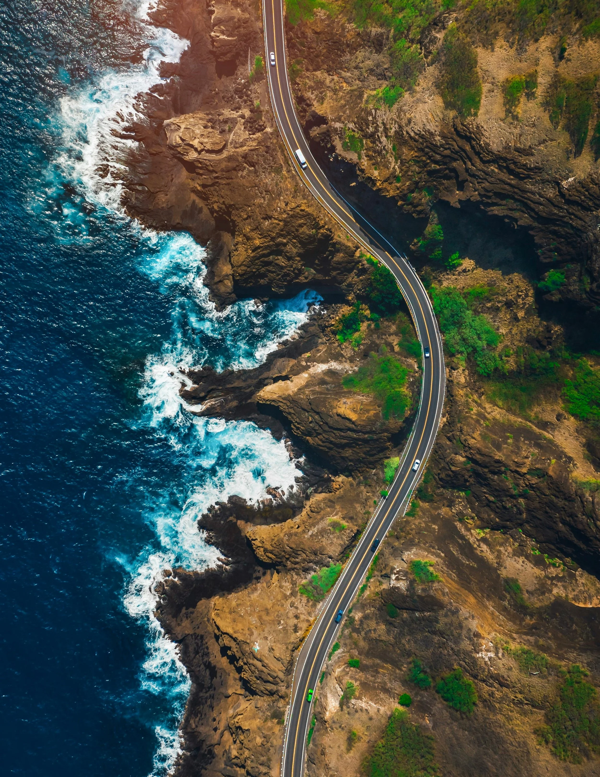 Aerial view of a coastal road winding along rugged cliffs with ocean waves crashing against rocks.