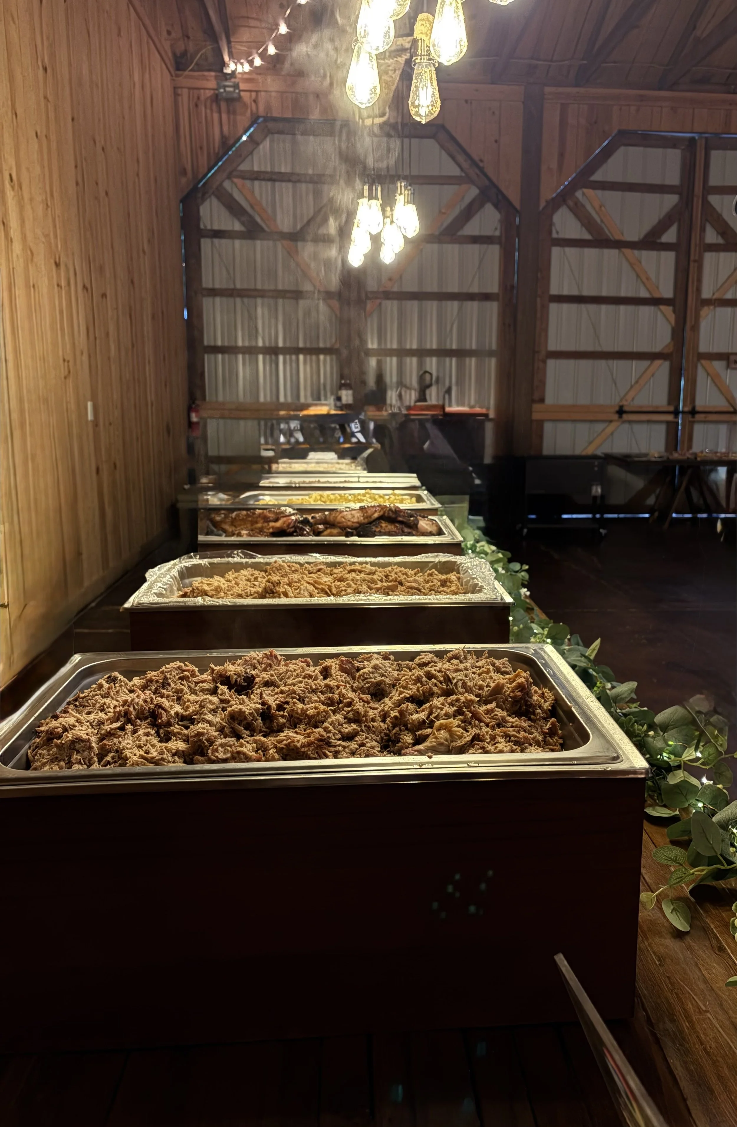 Buffet table with trays of shredded meat, cooked chicken, mashed potatoes, and corn, set in a rustic wooden barn with string lights and a chandelier.