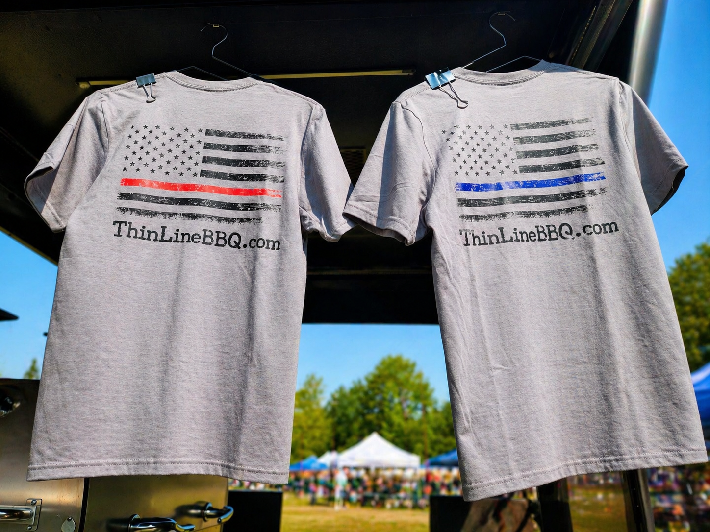 Two gray T-shirts hanging from hooks, featuring American flag designs with the stripes in distress style, one with a red stripe and the other with a blue stripe. The shirts display the website "ThinLineBBQ.com" below the flags, with a blurry outdoor 