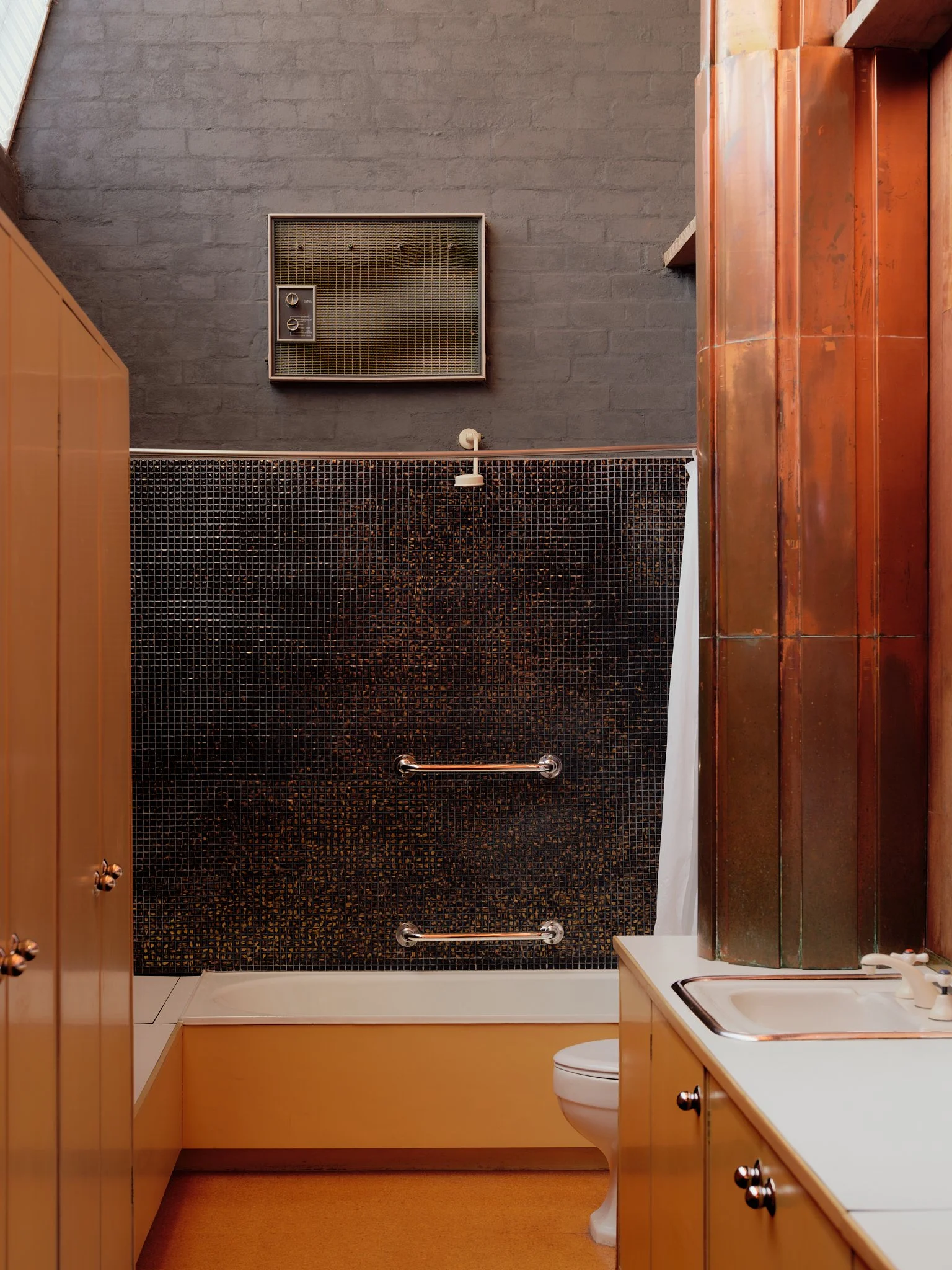 A small bathroom with brown cabinetry, a white toilet, a sink with a faucet, a dark tiled shower area with grab bars, and an industrial-style vent on the gray brick wall.