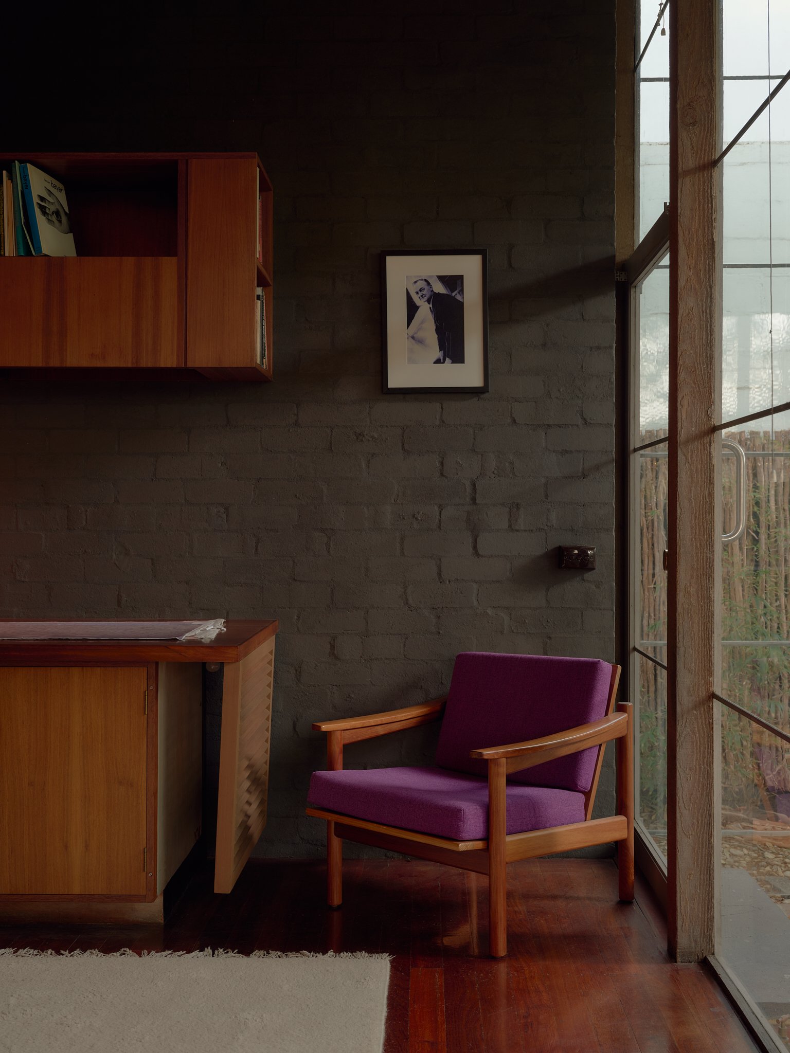 A cozy interior corner with a purple armchair next to a large glass door, a wooden desk, a wall-mounted wooden shelf with books, a black and white framed photo on a dark brick wall, and a white rug on polished wooden floor.
