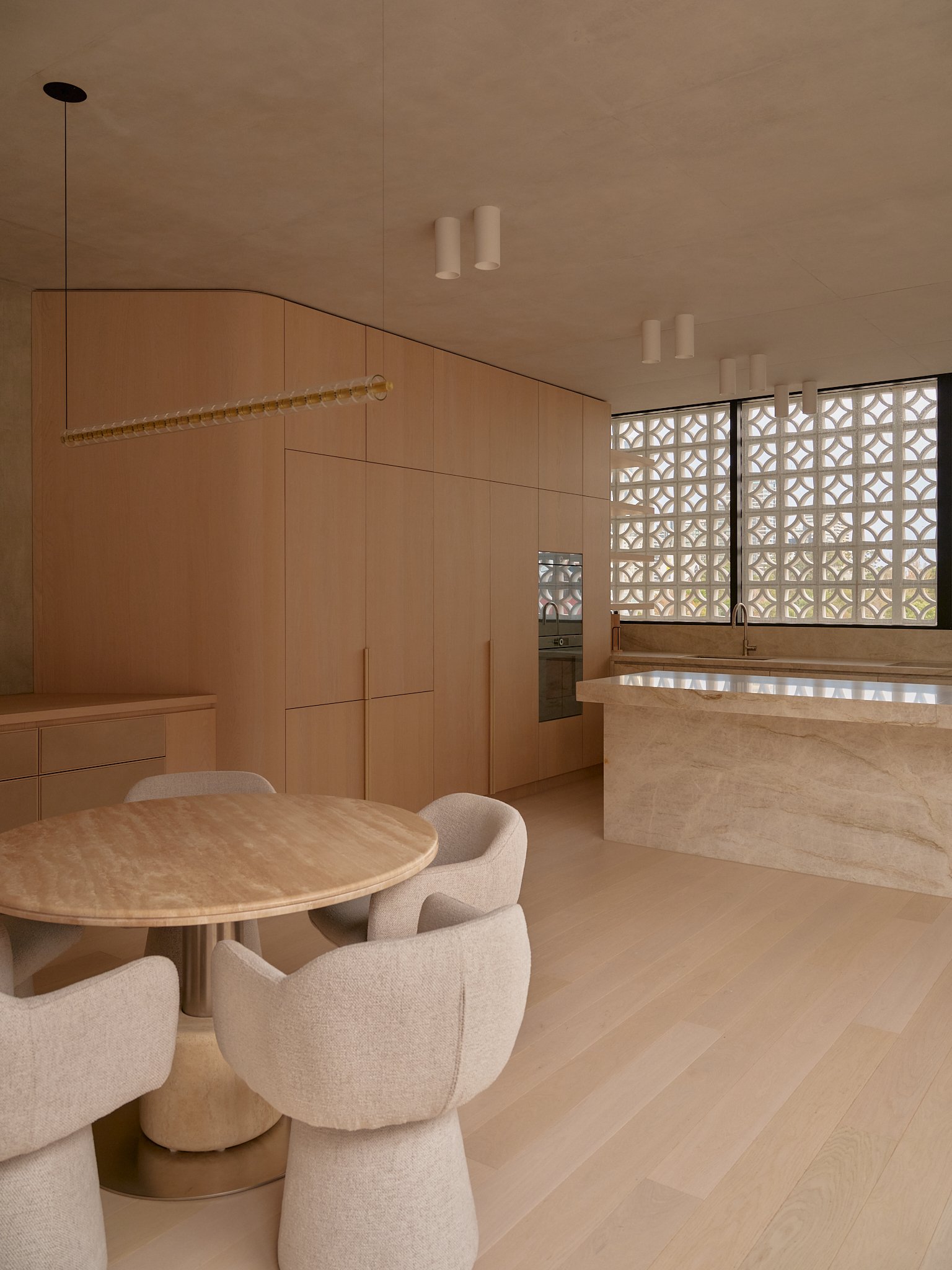 Modern kitchen with wooden cabinets, a marble island, a round dining table with six upholstered chairs, large windows with decorative privacy screen, and ceiling lights.