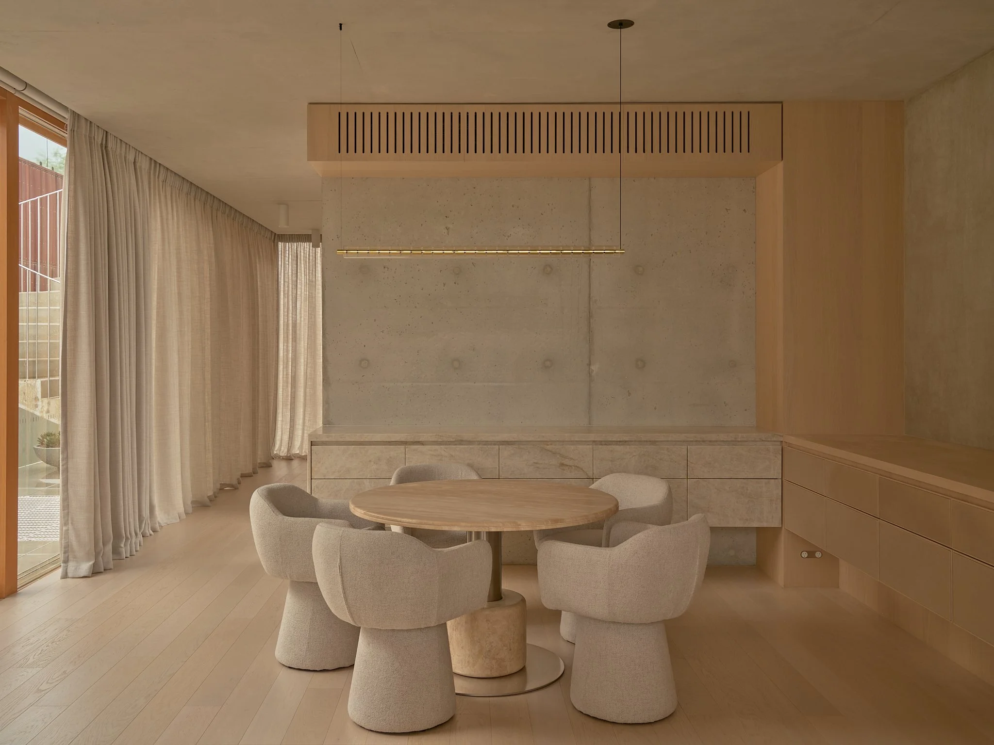 Minimalist dining area with round wooden table, six beige upholstered chairs, concrete wall, cream curtains, wooden flooring, and modern pendant lighting.