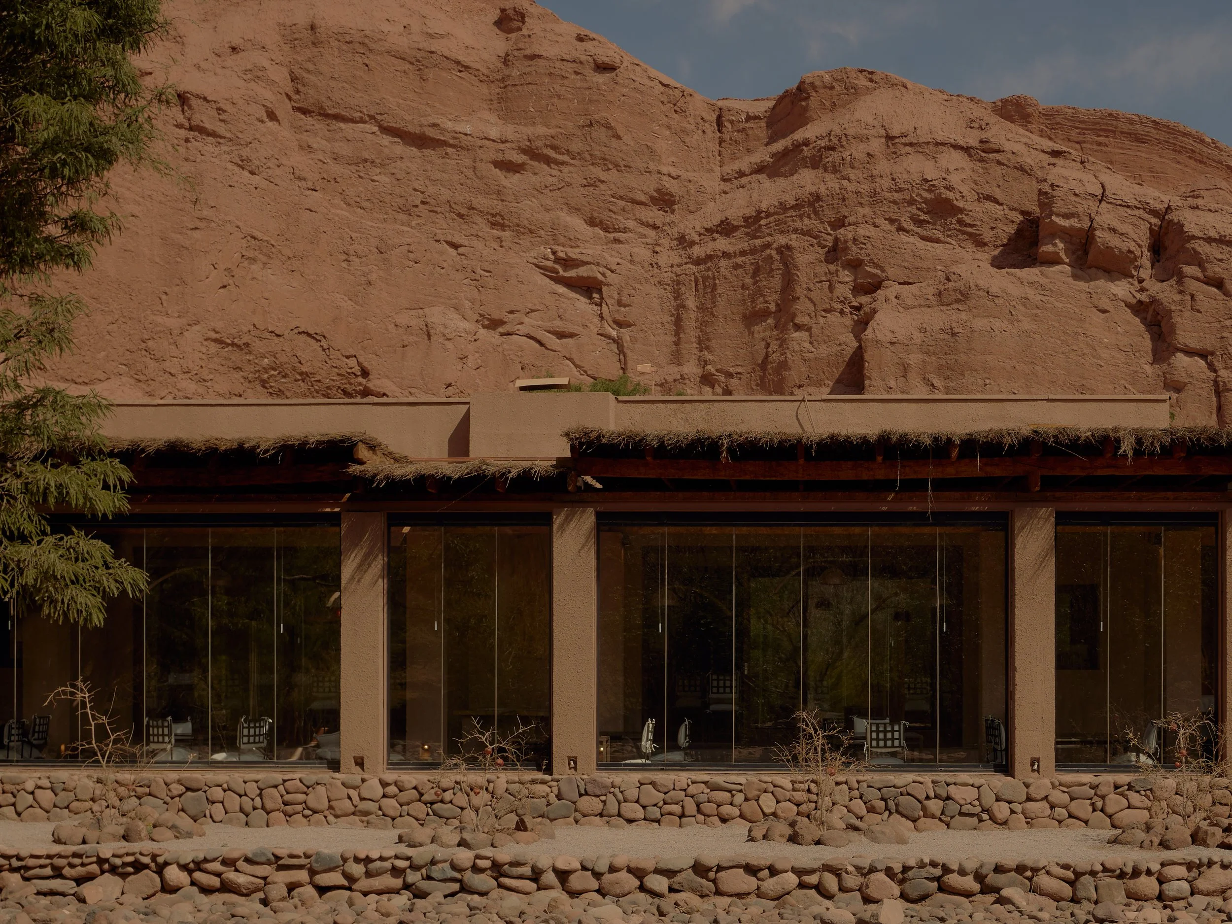 A modern building with glass walls situated at the base of a large, reddish-brown rocky cliff, with minimal landscaping featuring small rocks and sparse plants in the foreground.