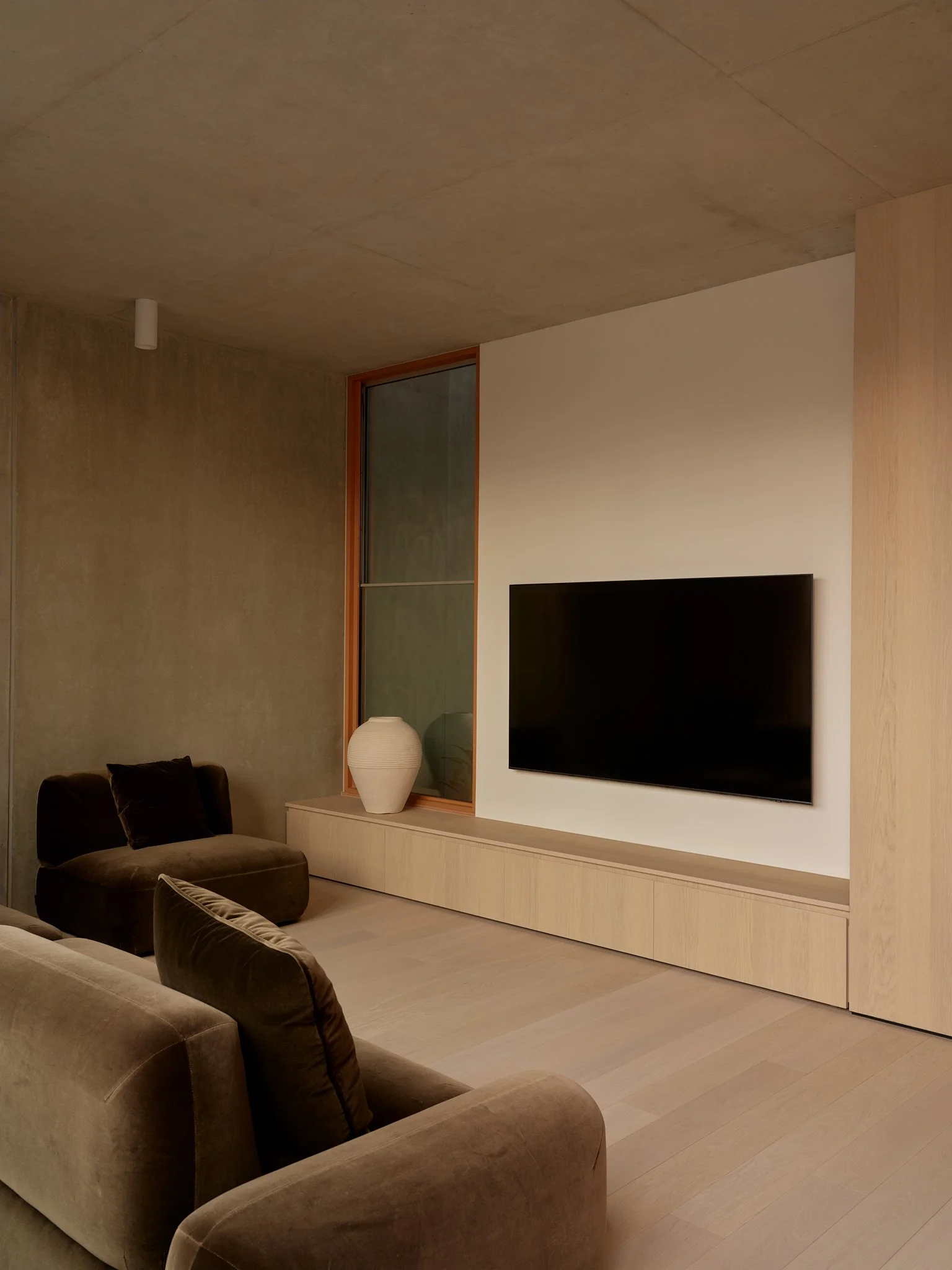 Modern living room with beige couches, a built-in wooden entertainment center, a large flat screen TV, a decorative white vase on the console, and a tall window.