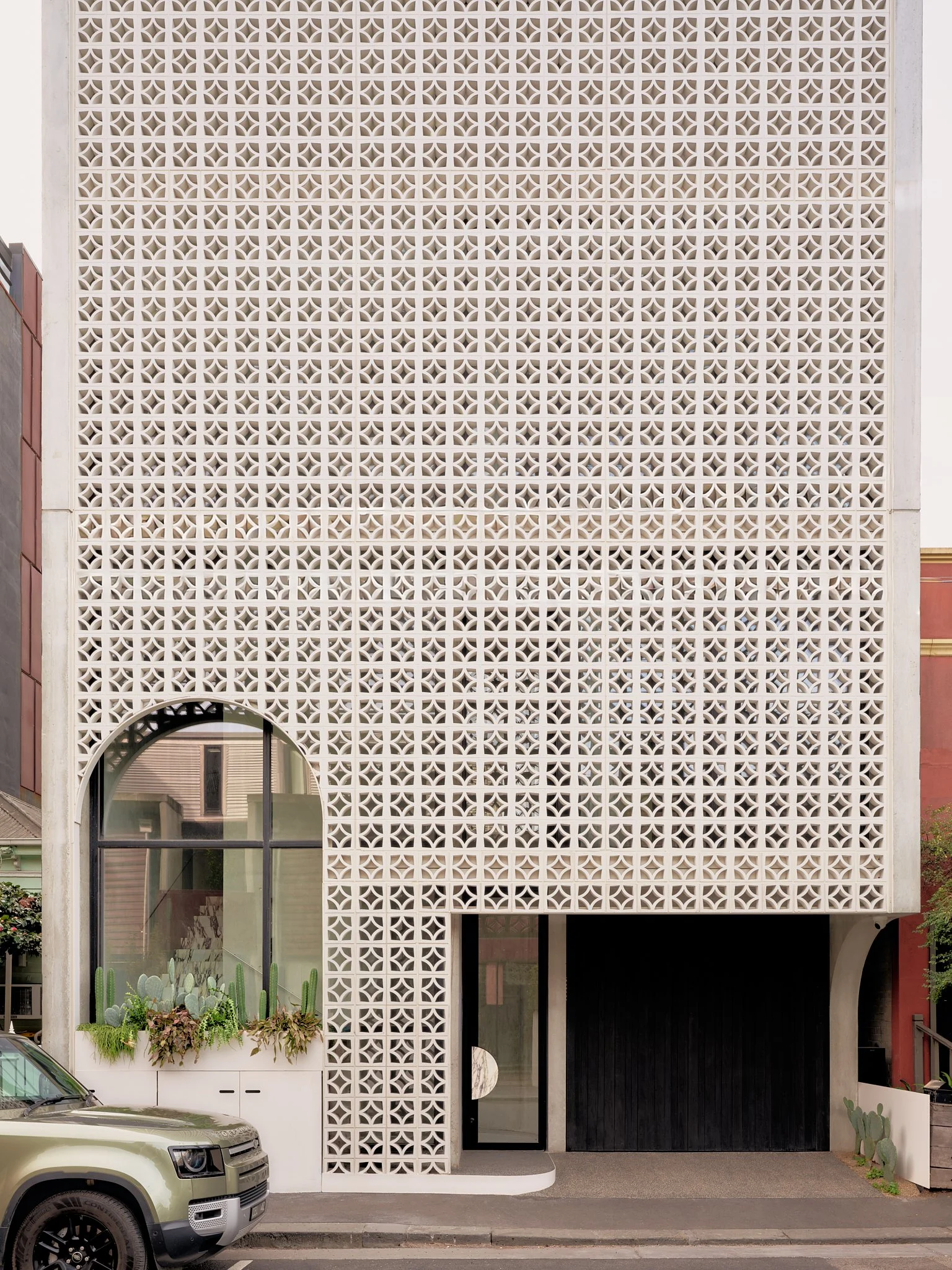 Modern building with a decorative white perforated facade, large arched window with plants inside, and a black door, parked car in front.