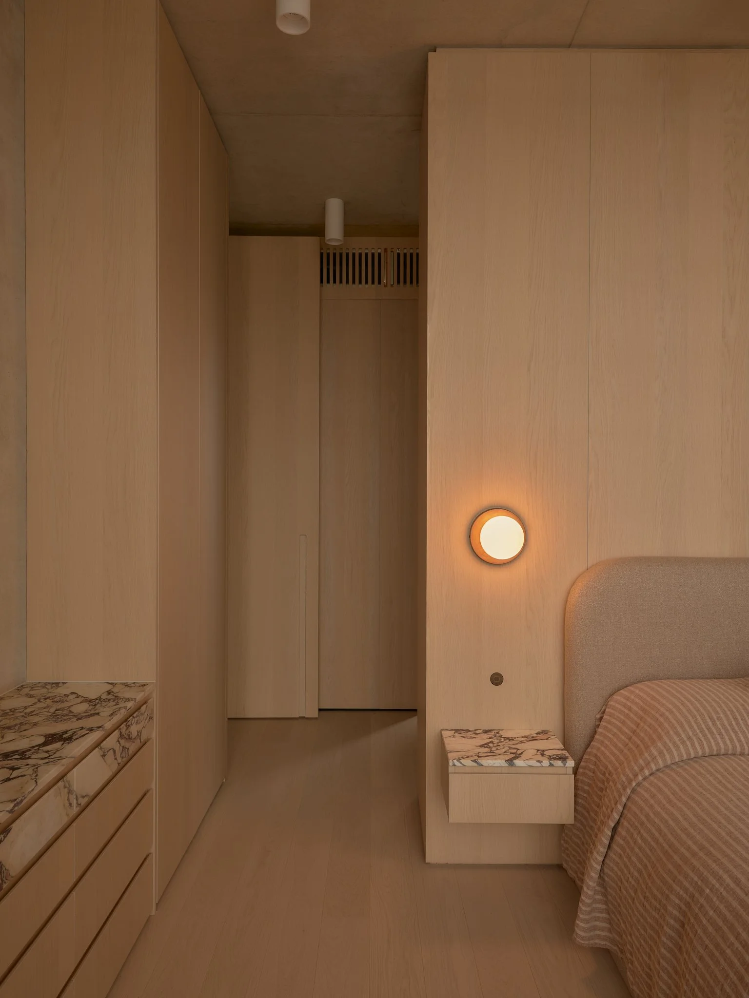A bedroom with light wood walls and furniture, a beige bed with a striped blanket, a small marble-topped nightstand, a round wall light, and a door at the end of the room.