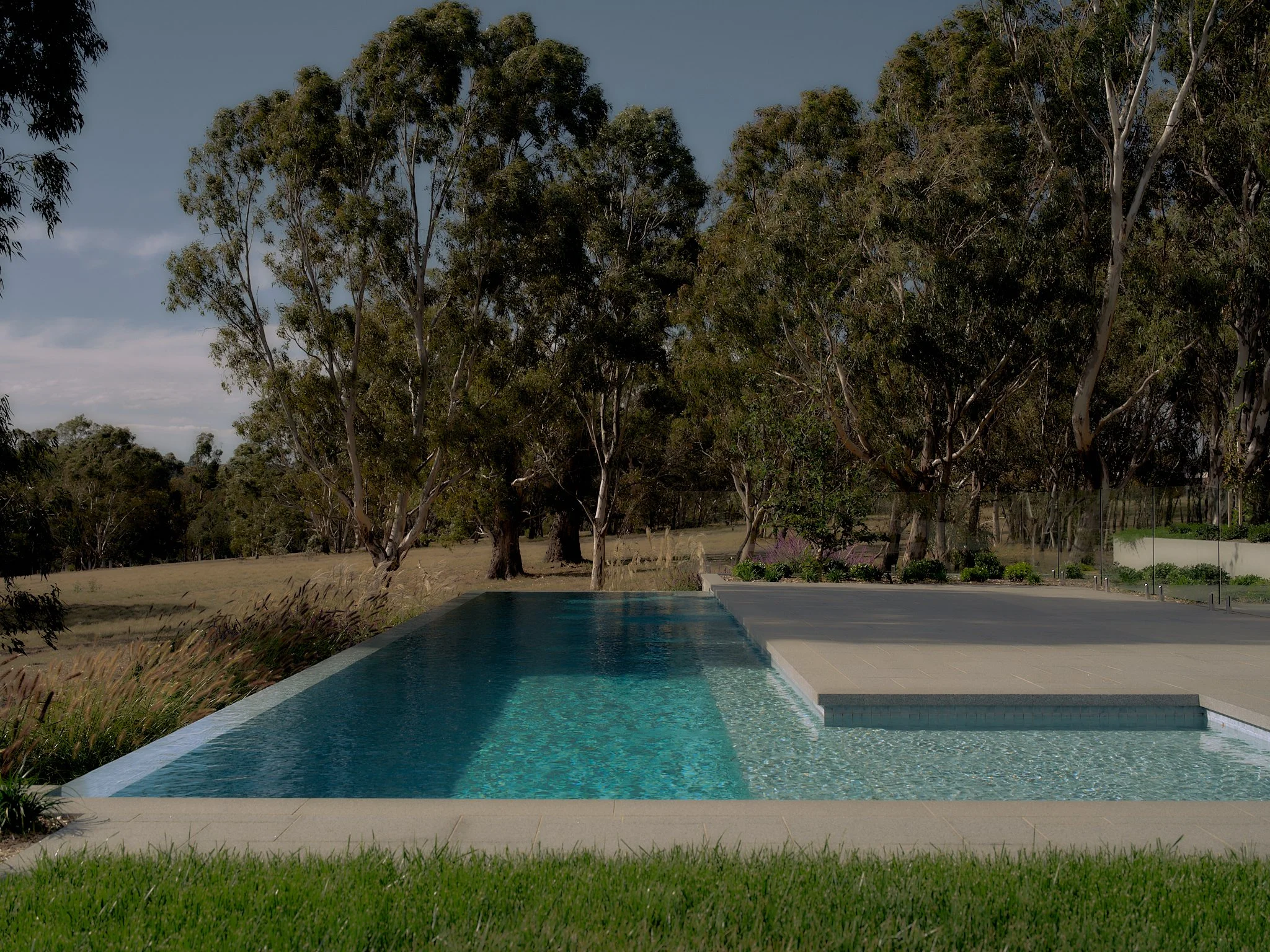 Modern infinity pool with clear water, adjacent to a gray stone patio, overlooking a grassy field and trees in the background on a partly cloudy day.