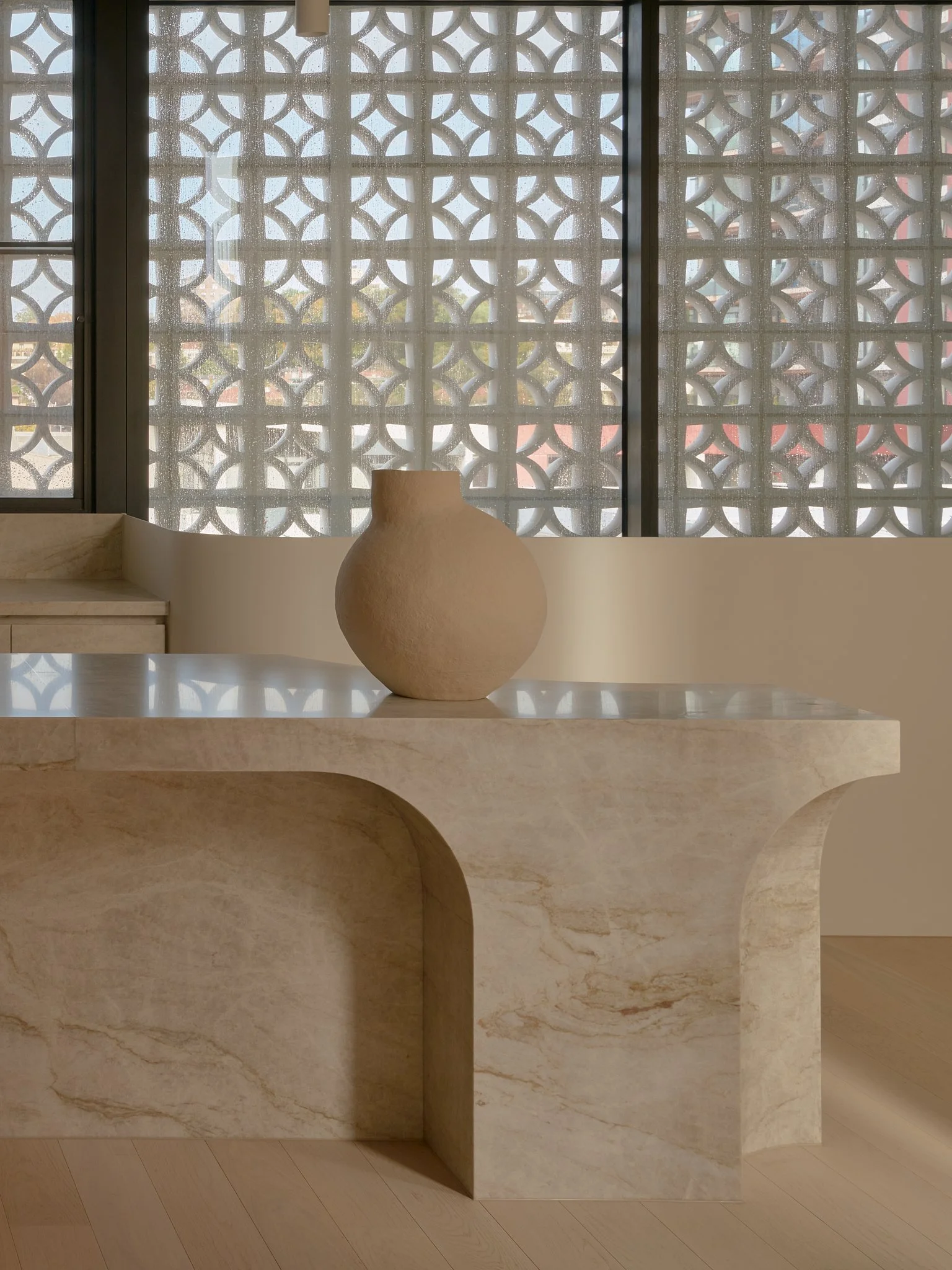 A beige, rounded vase on a pale, marble table with a curved edge, in front of a decorative screen with geometric patterns.
