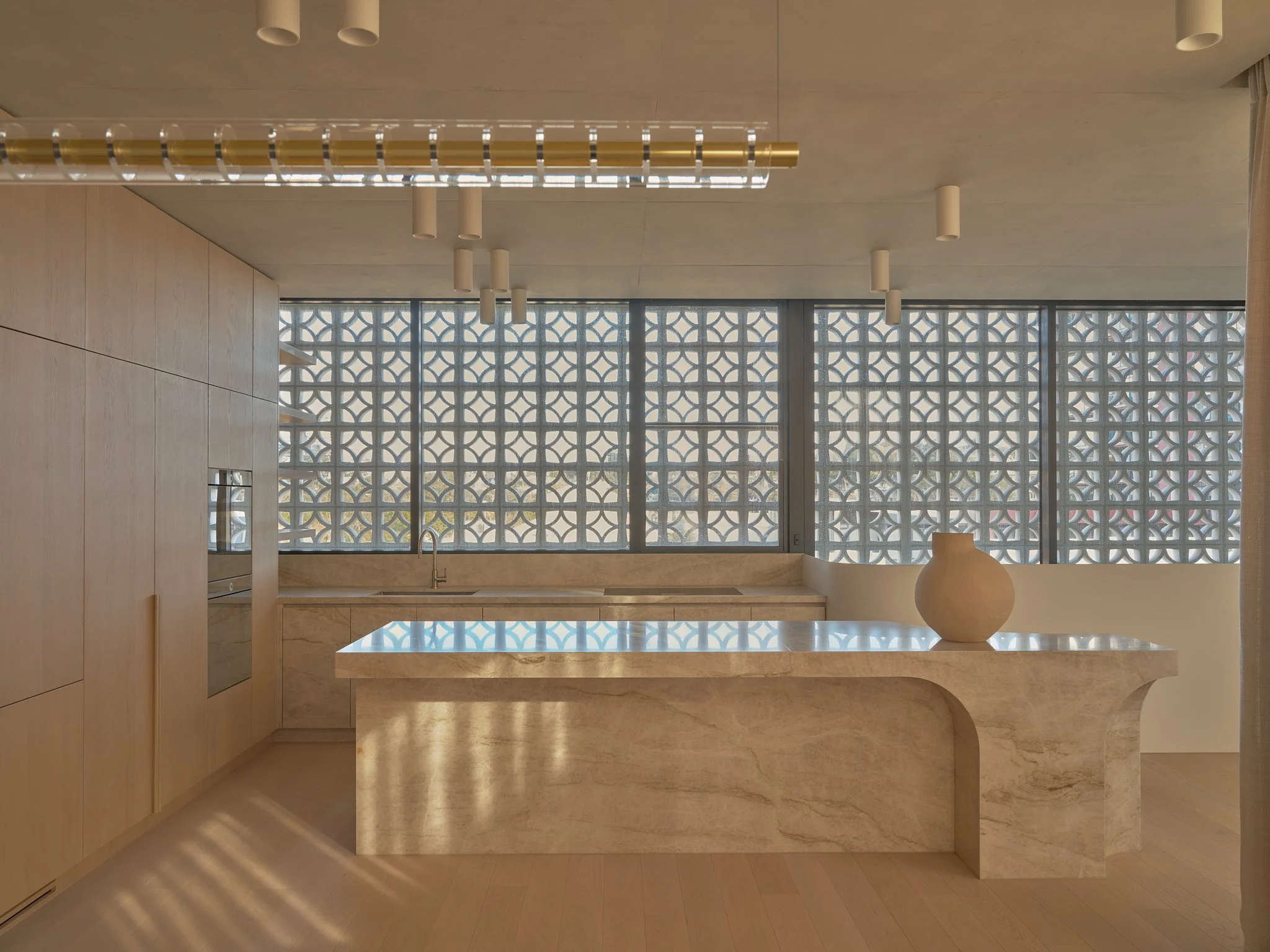 Modern kitchen with beige marble island, built-in oven, wooden cabinets, large window with decorative grille, and minimalist ceiling lighting.