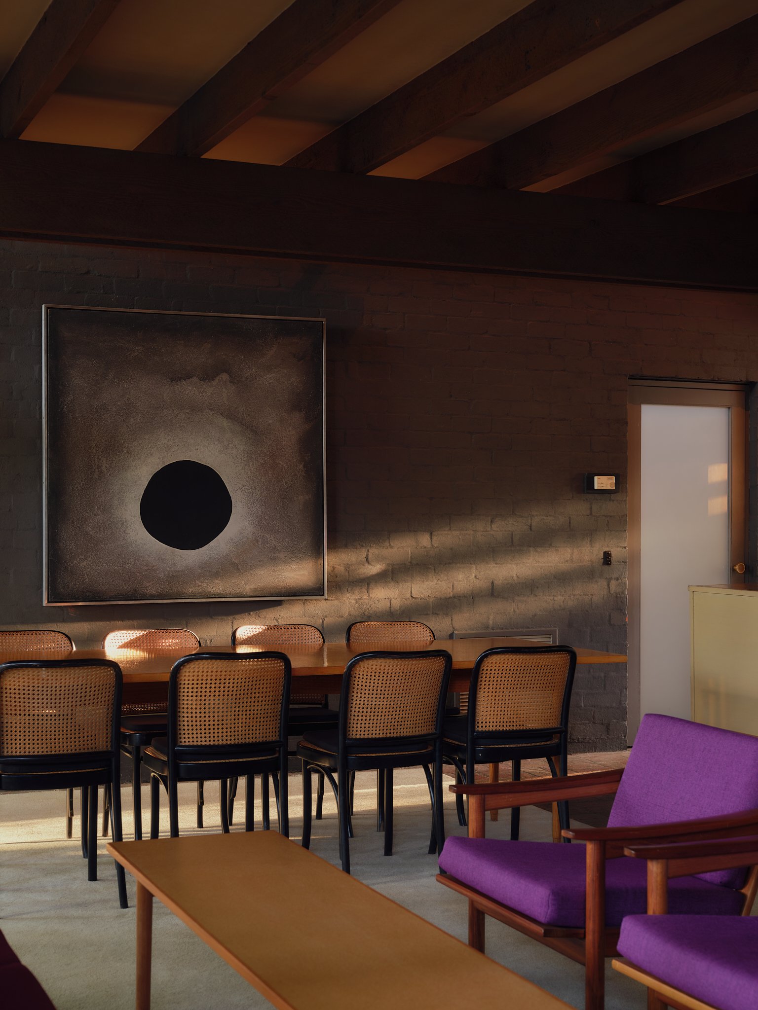 Interior of a modern room featuring a wooden table with six chairs, a purple armchair, abstract art on a brick wall, and sunlight casting shadows across the furniture.