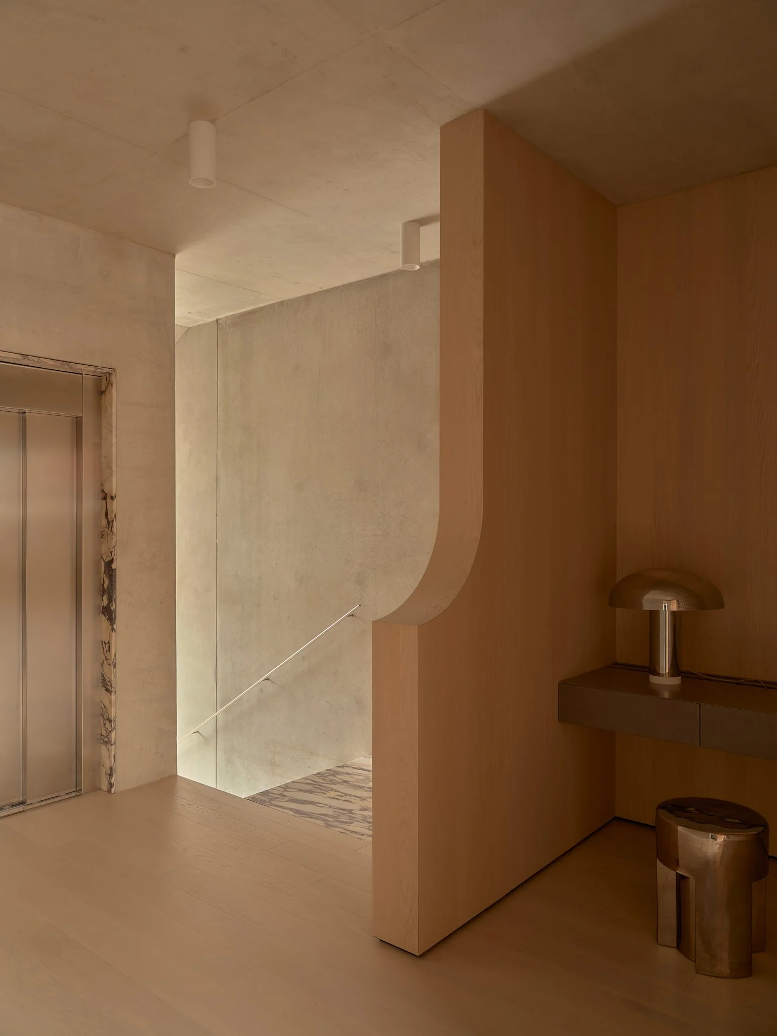 Minimalist interior with wooden wall, small table with modern lamp, elevator door, and a stairway visible down a corridor.