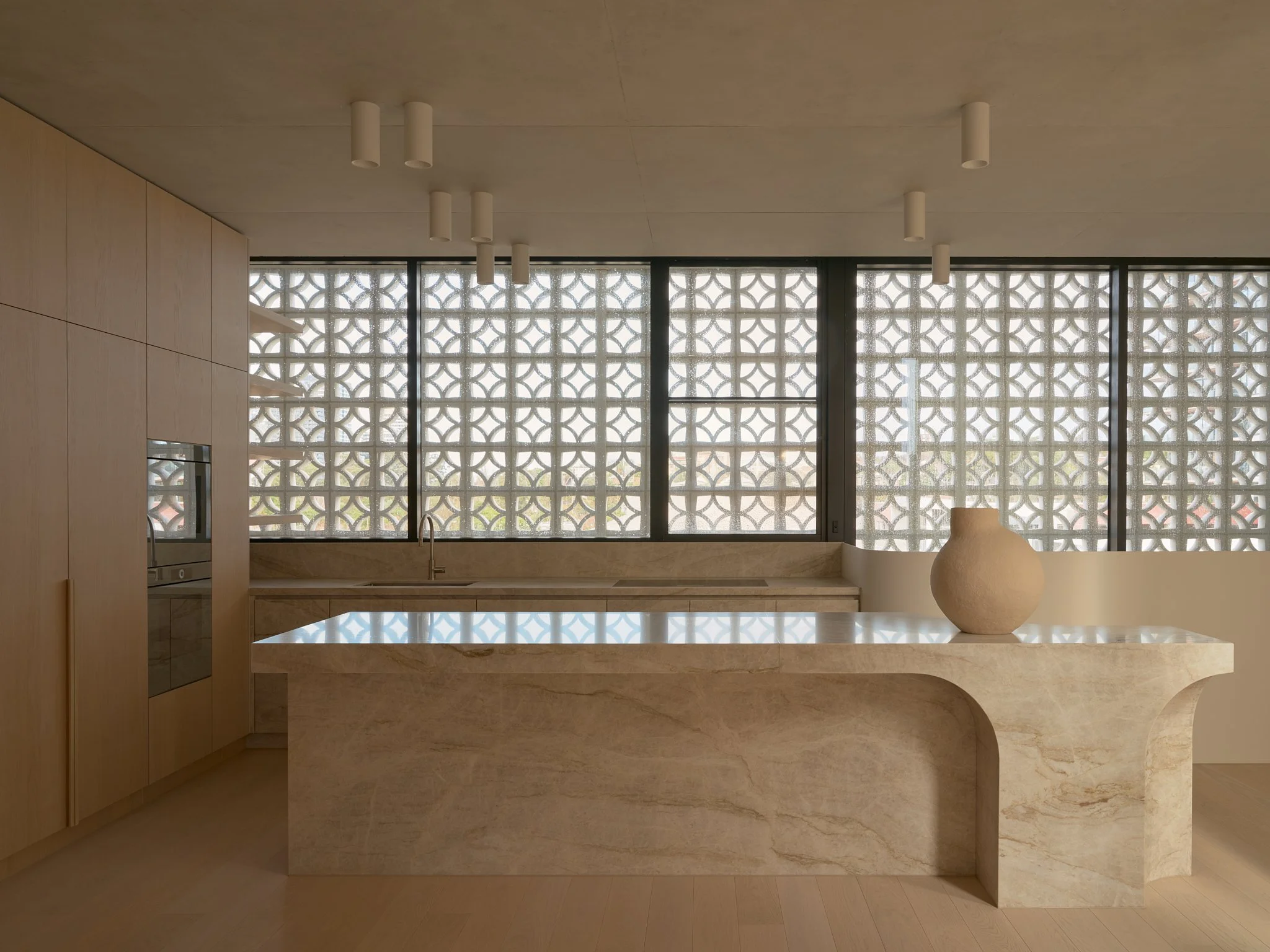 Minimalist kitchen with large patterned windows, light wood cabinets, marble island, and beige decorative vase.