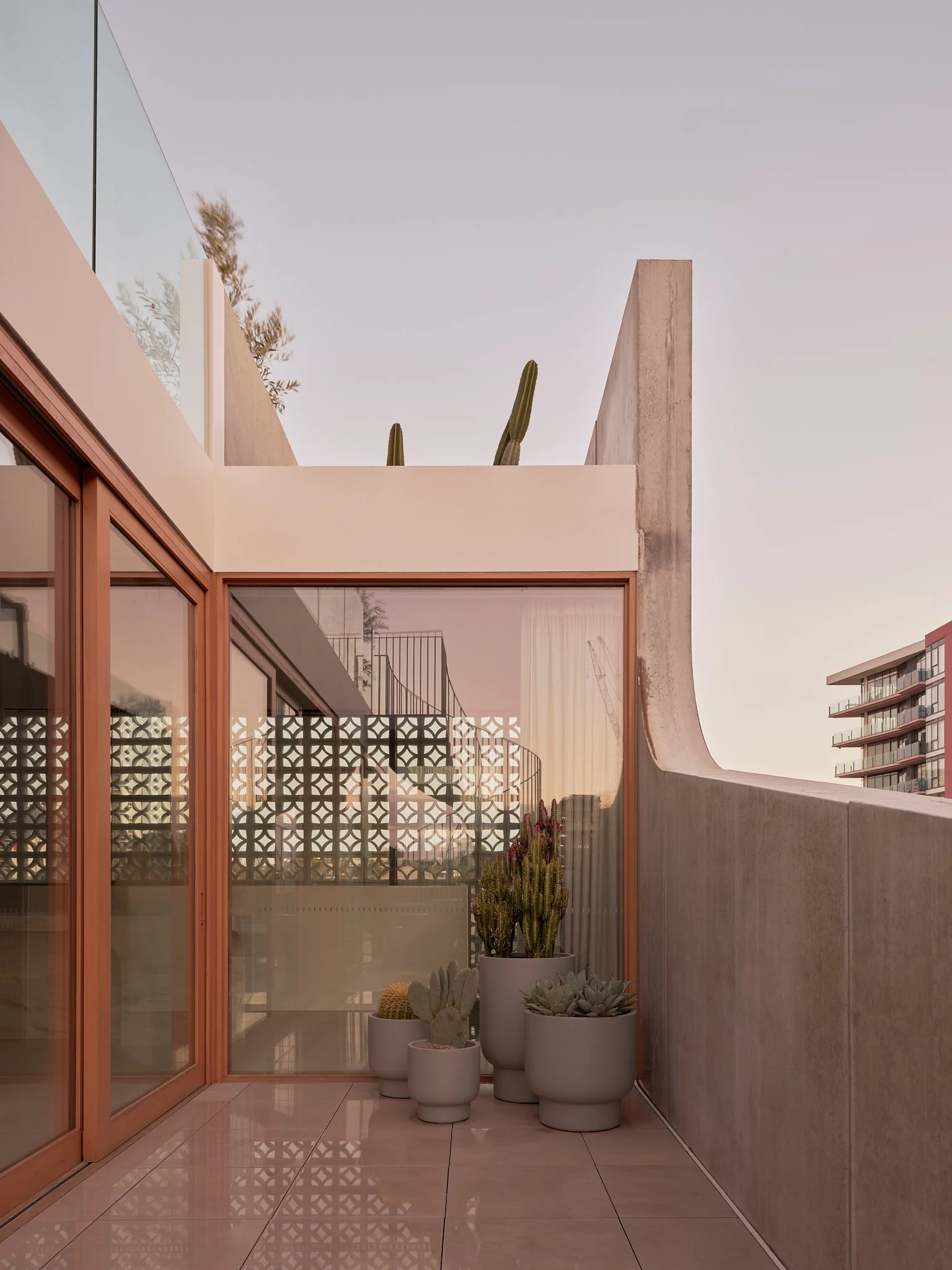 Modern balcony with potted cacti and succulents, glass sliding doors, and a decorative wall panel, overlooking a city skyline at dusk.