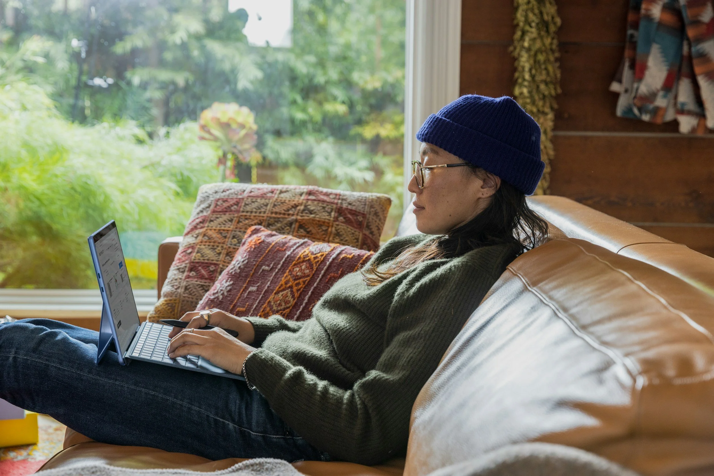 A person with glasses and a blue knit beanie is sitting on a beige couch, working on a small blue laptop, in a room with a large window showing a green outdoor scene.