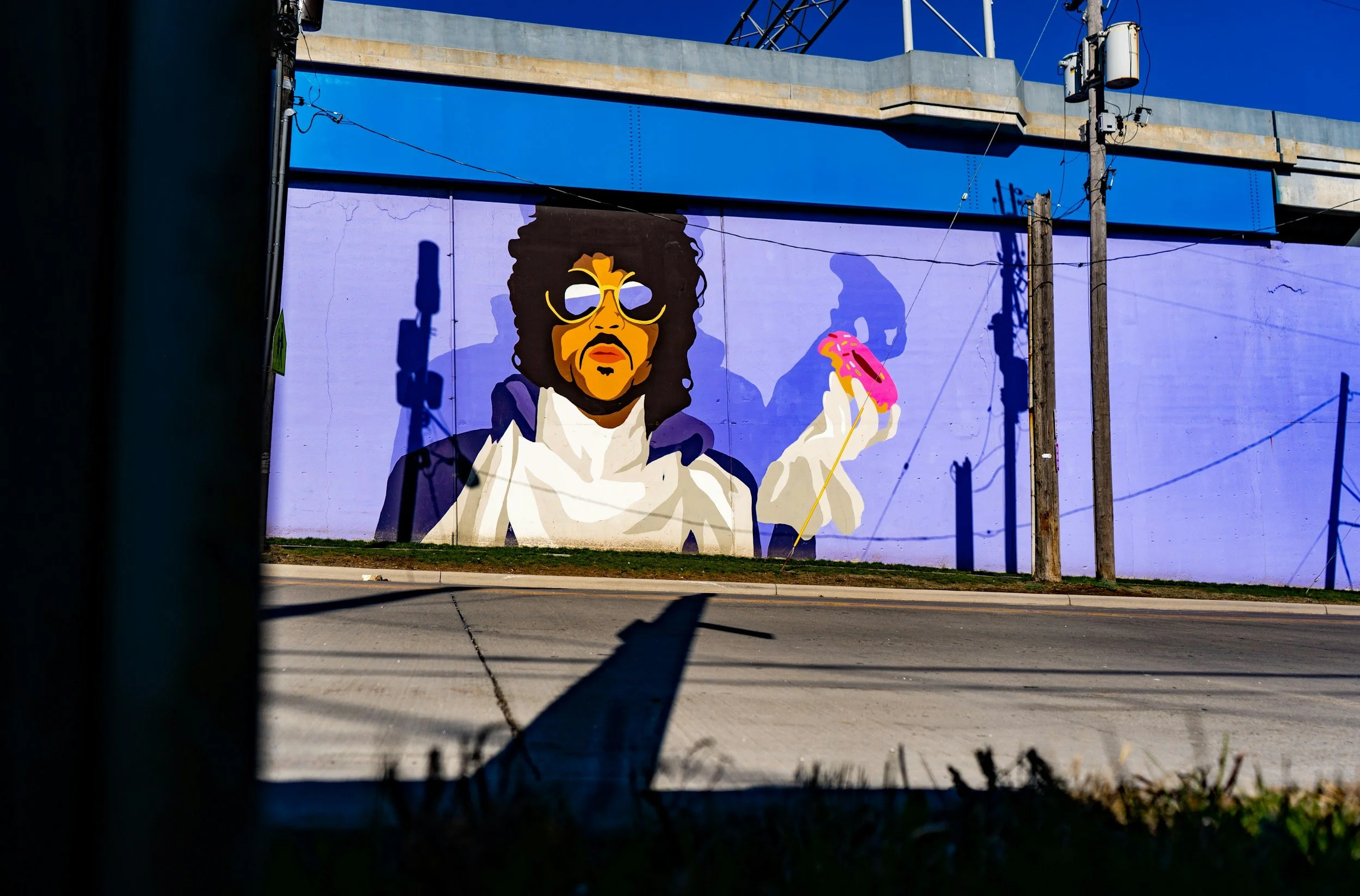 Street view with a colorful mural of a woman holding a pink donut on a blue building wall, seen through a gap with shadows and telephone poles.