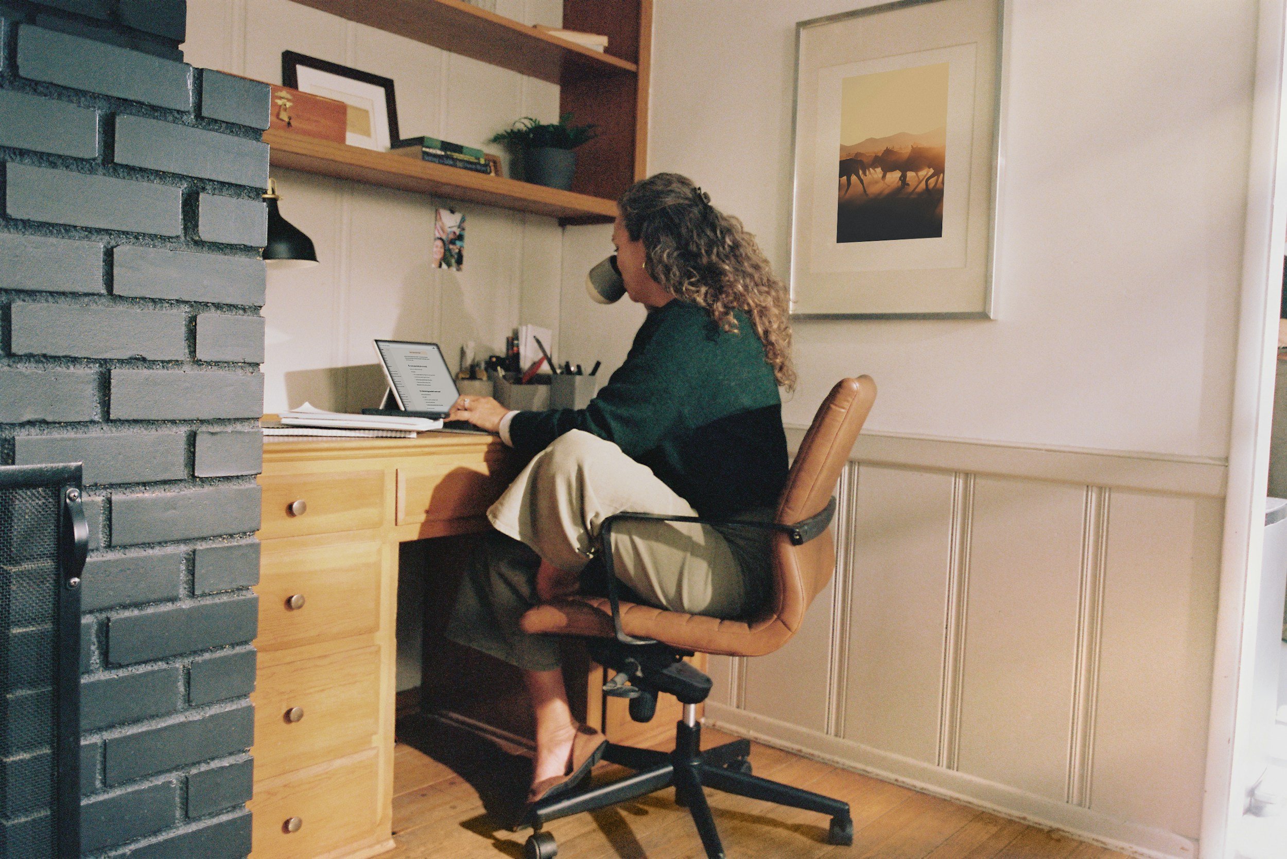 Woman sitting at a desk with a tablet, working or reading, in a cozy home office with framed art and shelves on the wall.