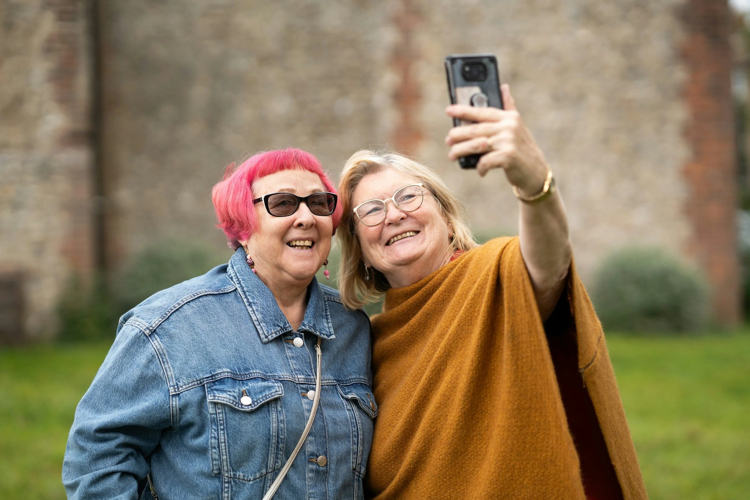 Two older women are taking a selfie outdoors. One has short pink hair and sunglasses, wearing a denim jacket. The other has light brown hair, glasses, and is wearing a mustard-colored sweater. They are smiling and standing close together.