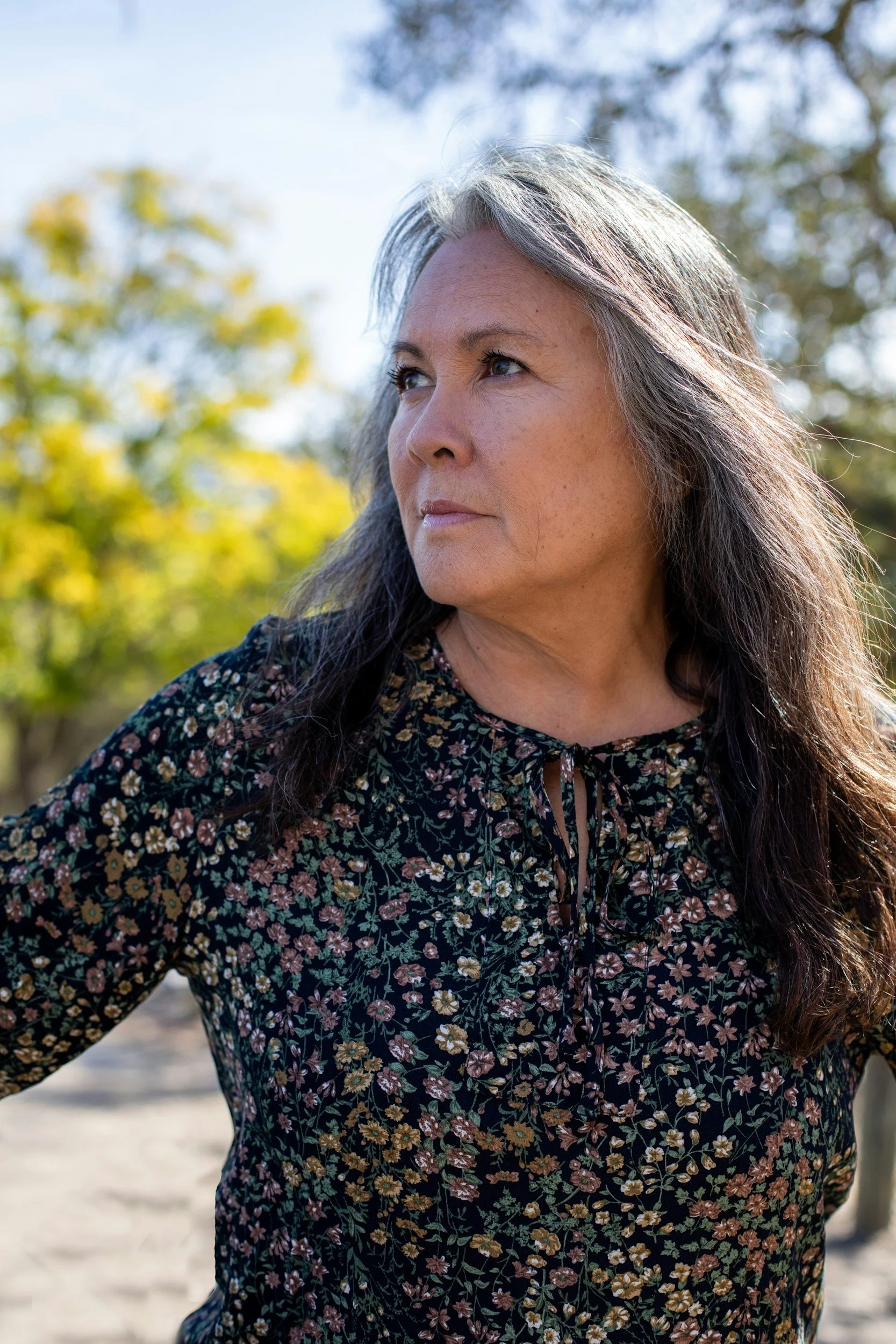 A woman with long gray hair and light skin, looking to her left outdoors on a sunny day with blurred trees and sky in the background, wearing a floral patterned dress.