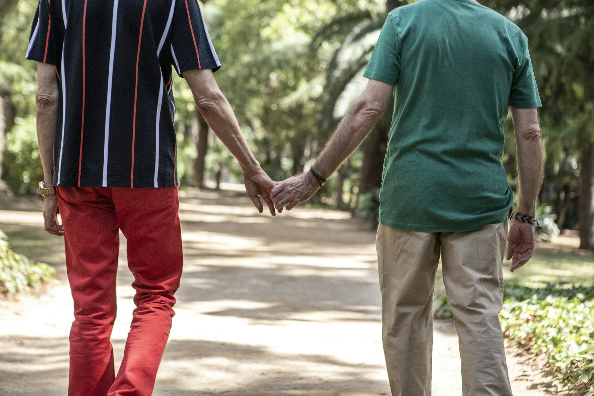 Two older people walking hand in hand outdoors along a tree-lined path on a sunny day.