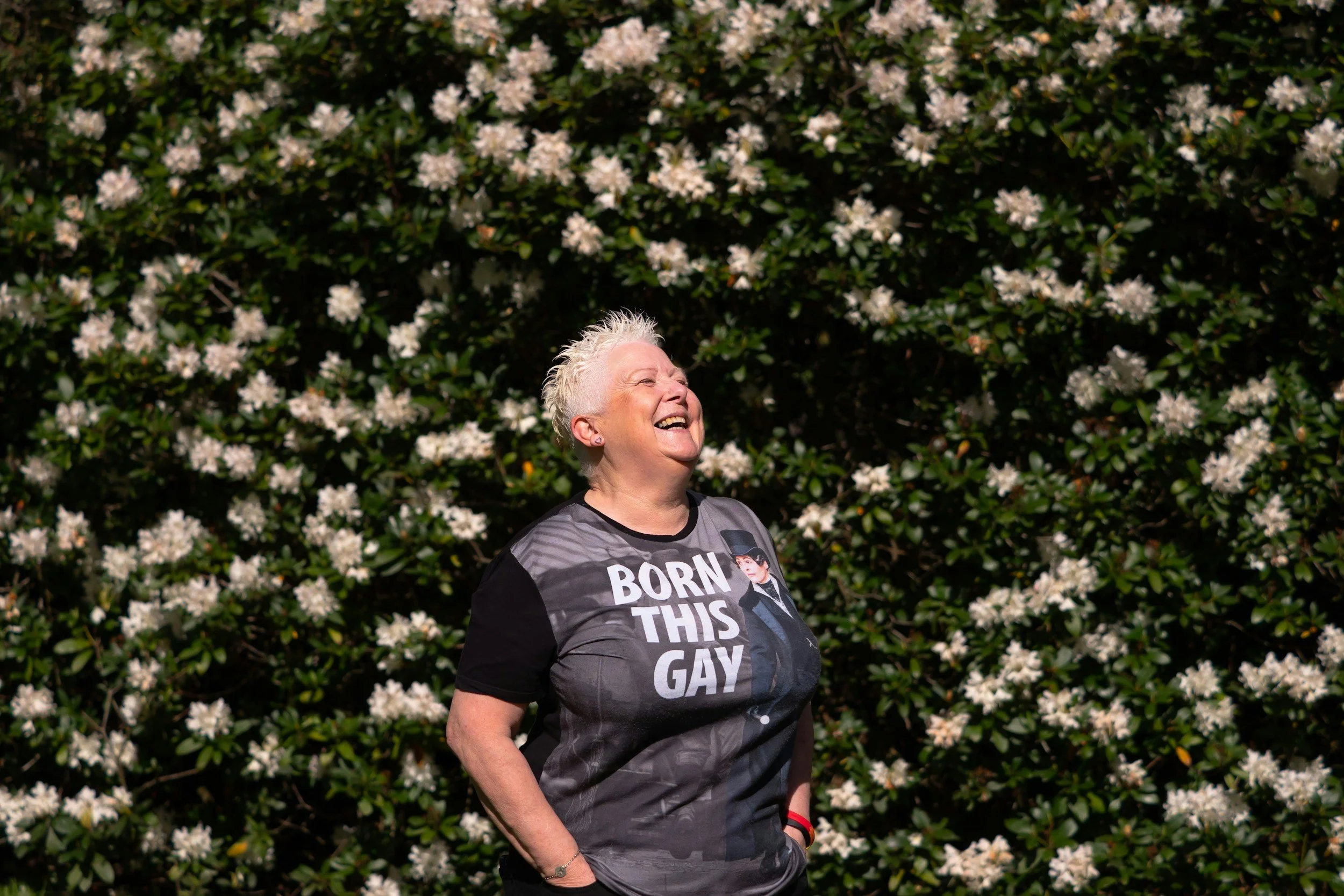 Smiling woman with short white hair wearing a graphic t-shirt reading 'Born This Gay,' standing outdoors in front of a bush with white flowers.