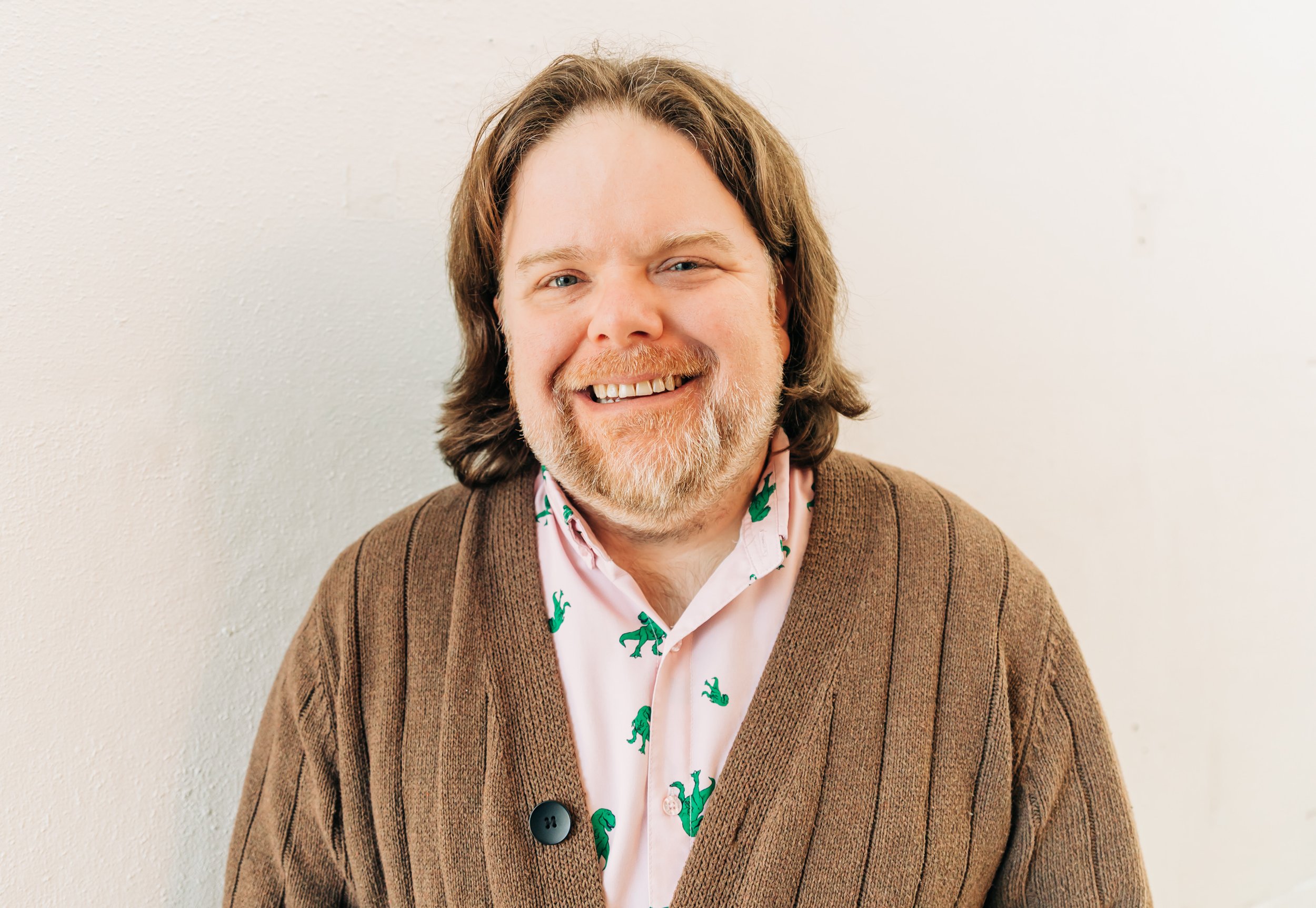 A smiling man with shoulder-length hair and a beard, wearing a brown sweater over a pink shirt with green dinosaur prints, standing against a plain white wall.