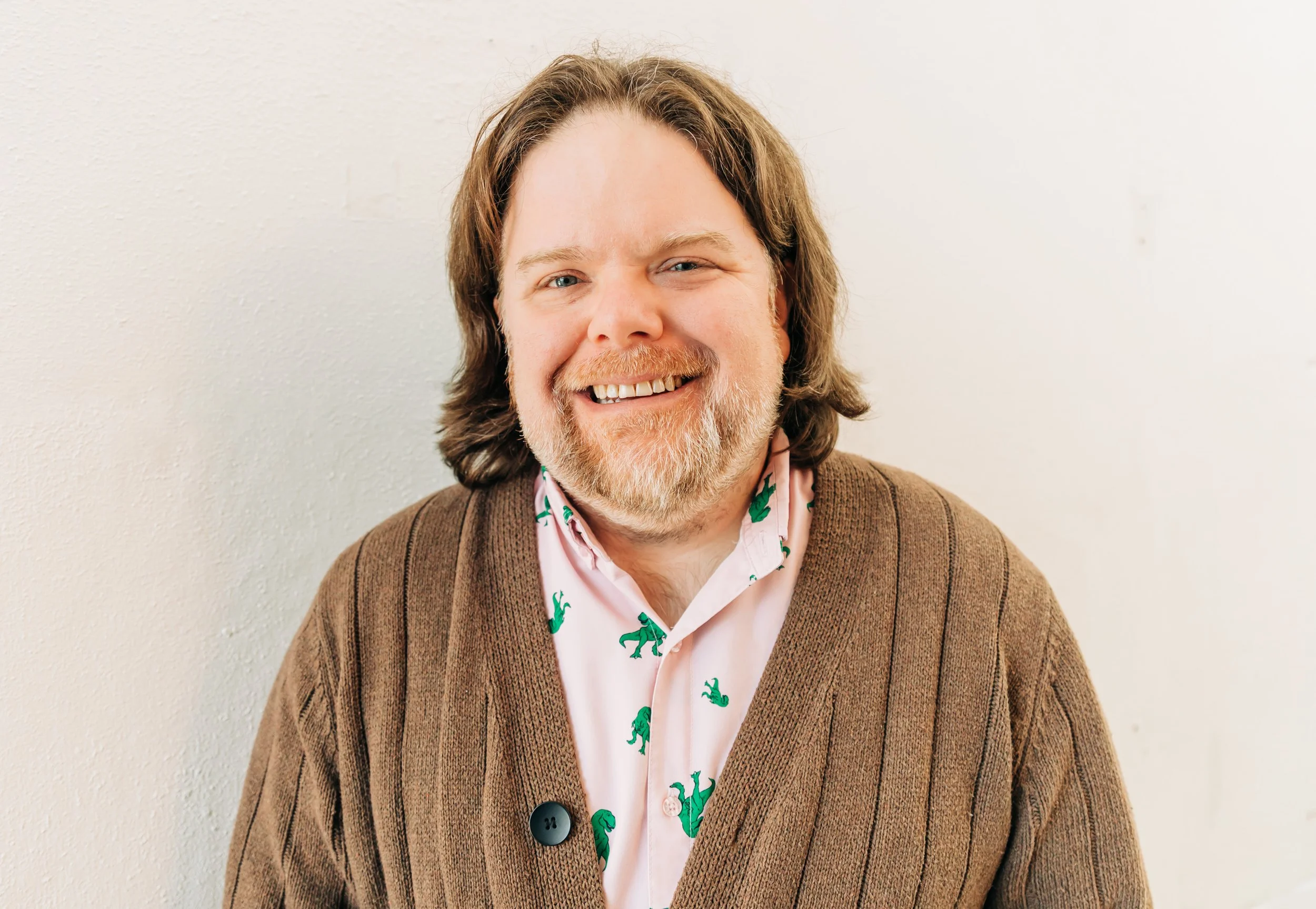 A smiling man with long brown hair, a beard, and glasses, wearing a pink shirt with green dinosaur prints and a brown cardigan, standing against a plain light-colored wall.