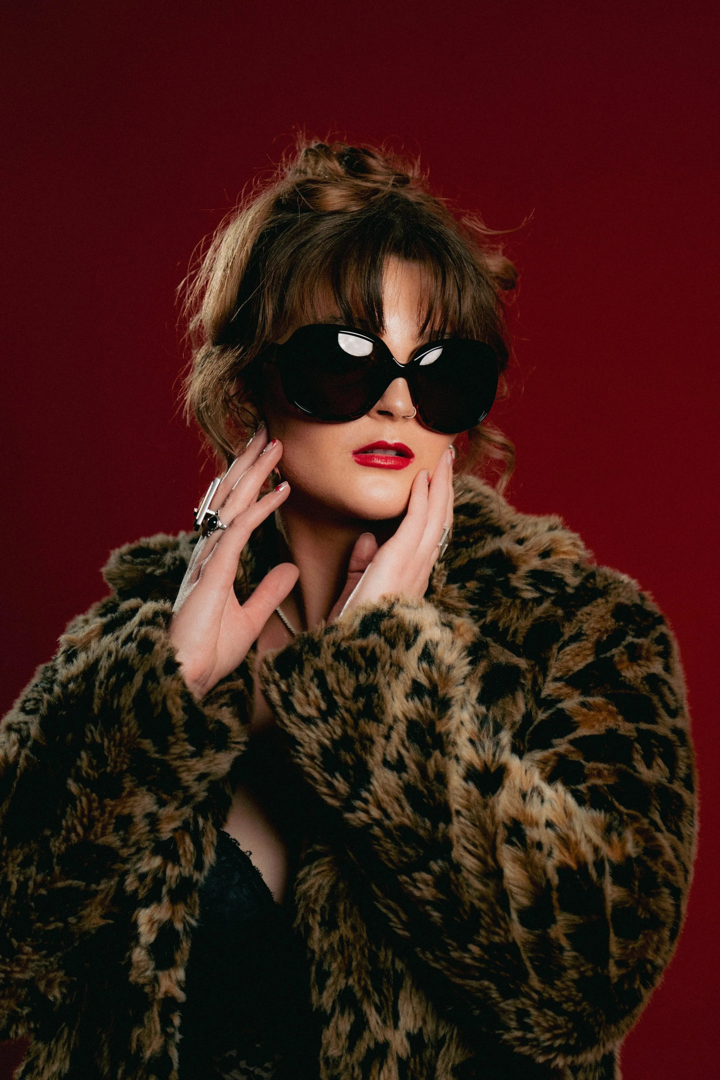 A woman with tousled, shoulder-length hair wearing large black sunglasses, a leopard-print fur coat, and red lipstick, posing against a red background.