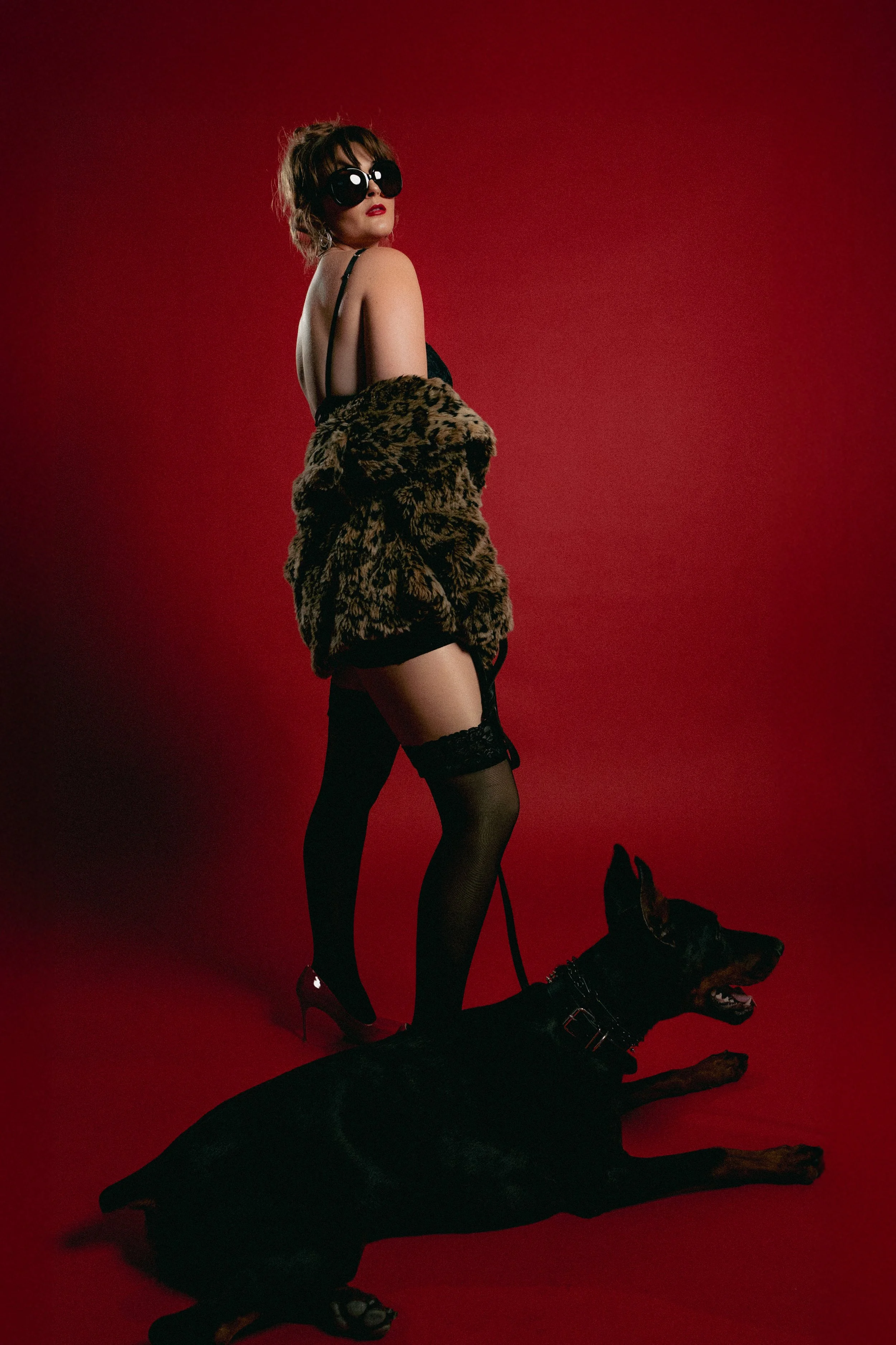 A woman with brown hair in a short curly hairstyle wearing large black sunglasses, a black bra, black thigh-high stockings, and high heels, stands with a black Doberman dog on a red background. The woman has a leopard print fur coat draped over her hips. The dog is laying on the ground with a leash held by the woman, and the woman's pose is turned to the side with her head turned towards the camera.