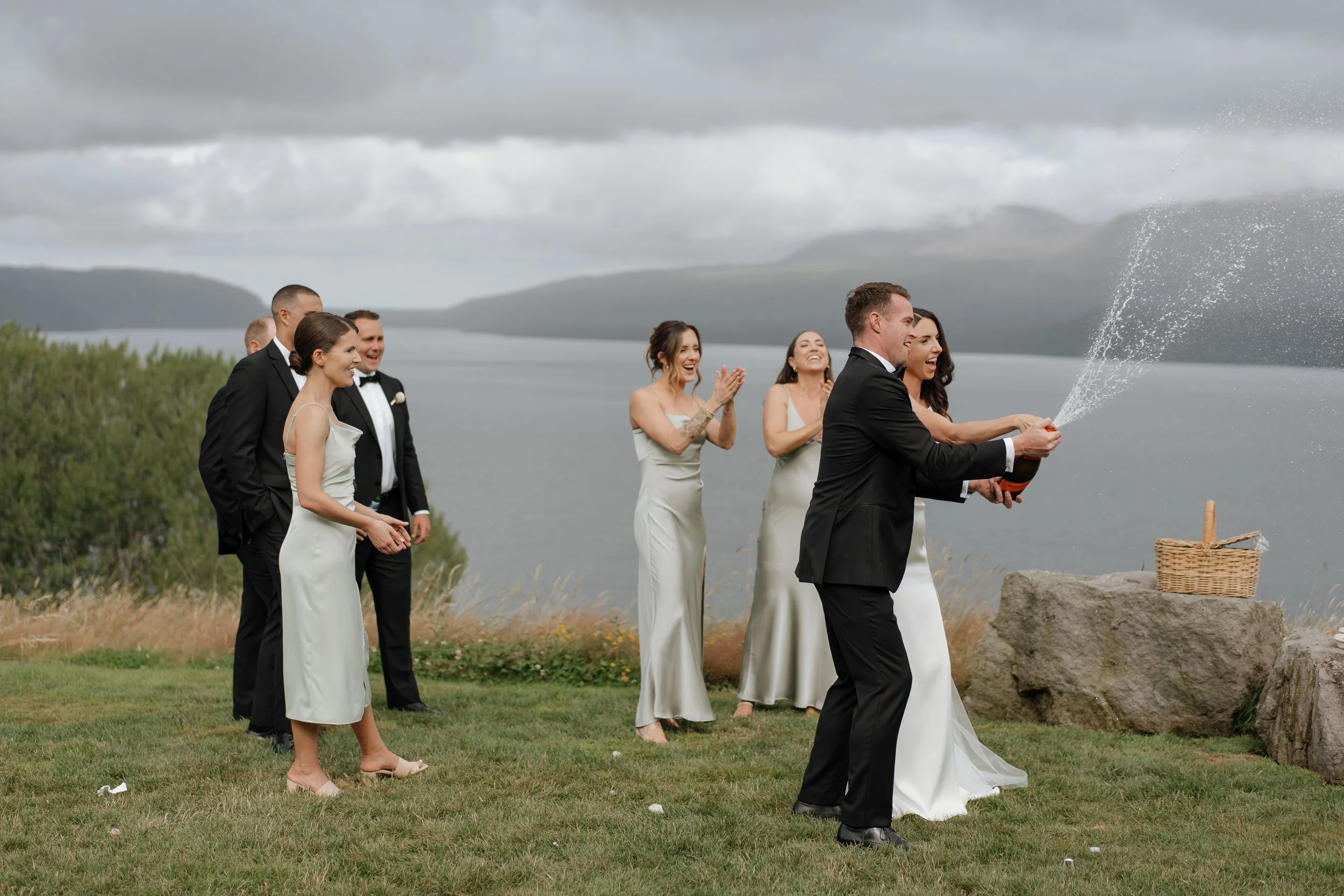 Wedding celebration outdoors by a lake on a cloudy day, featuring a group of people in formal attire, with a woman in a wedding dress and a man in a tuxedo popping a champagne bottle, while others cheer.