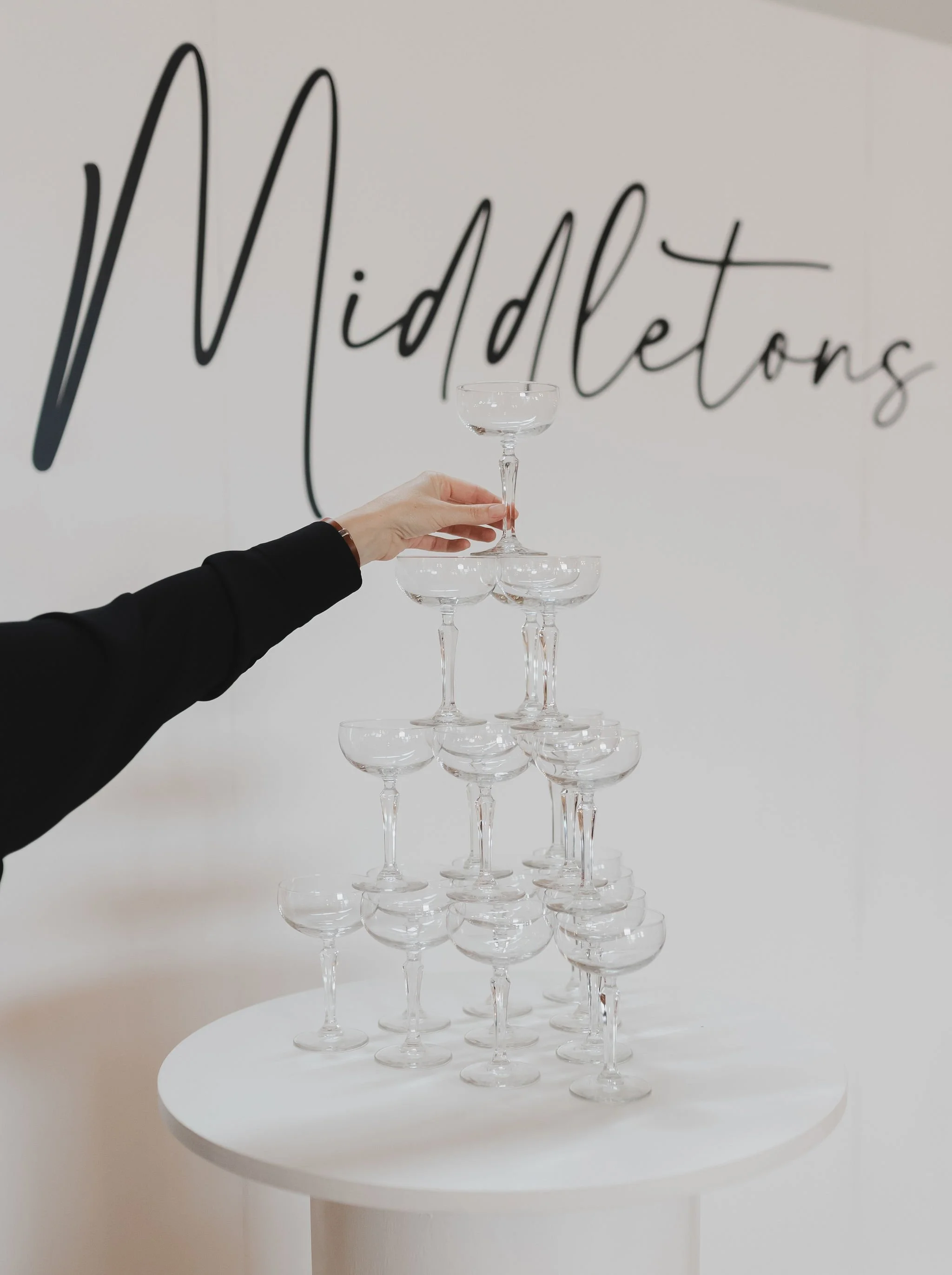 A person stacking champagne flutes on a white table in front of a wall with black cursive text that reads 'Midleton's'.