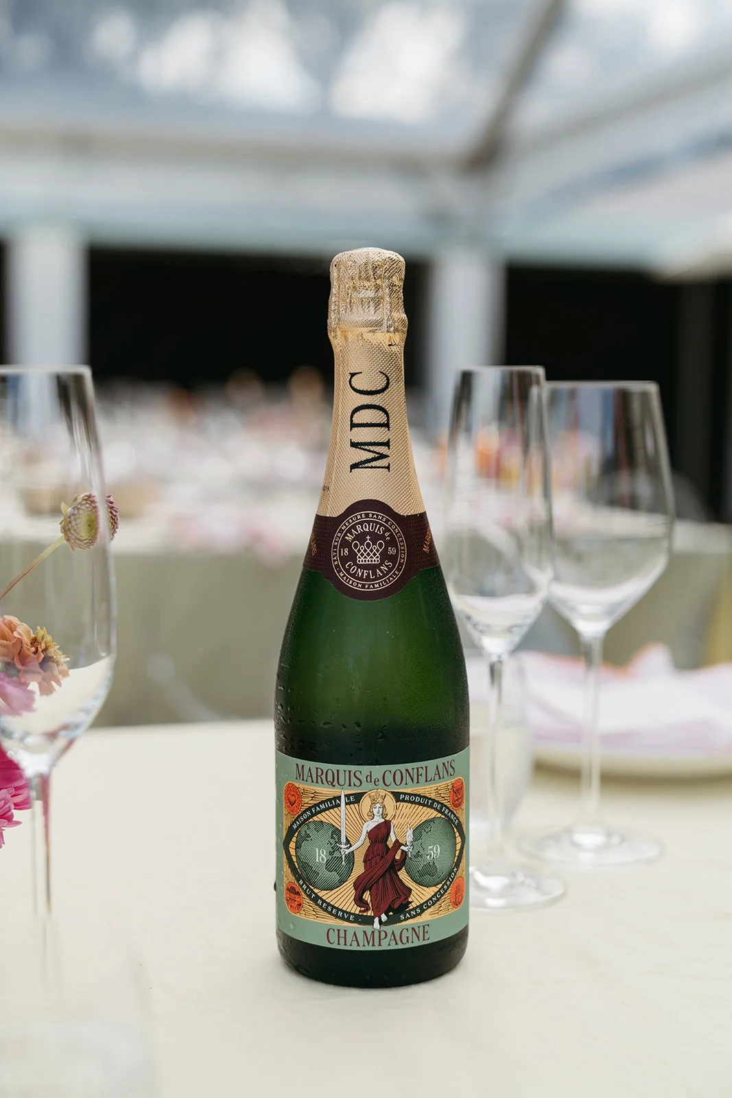 A bottle of Marquis de Conflans Champagne on a white table, surrounded by empty wine glasses and pink flowers in a glass vase.