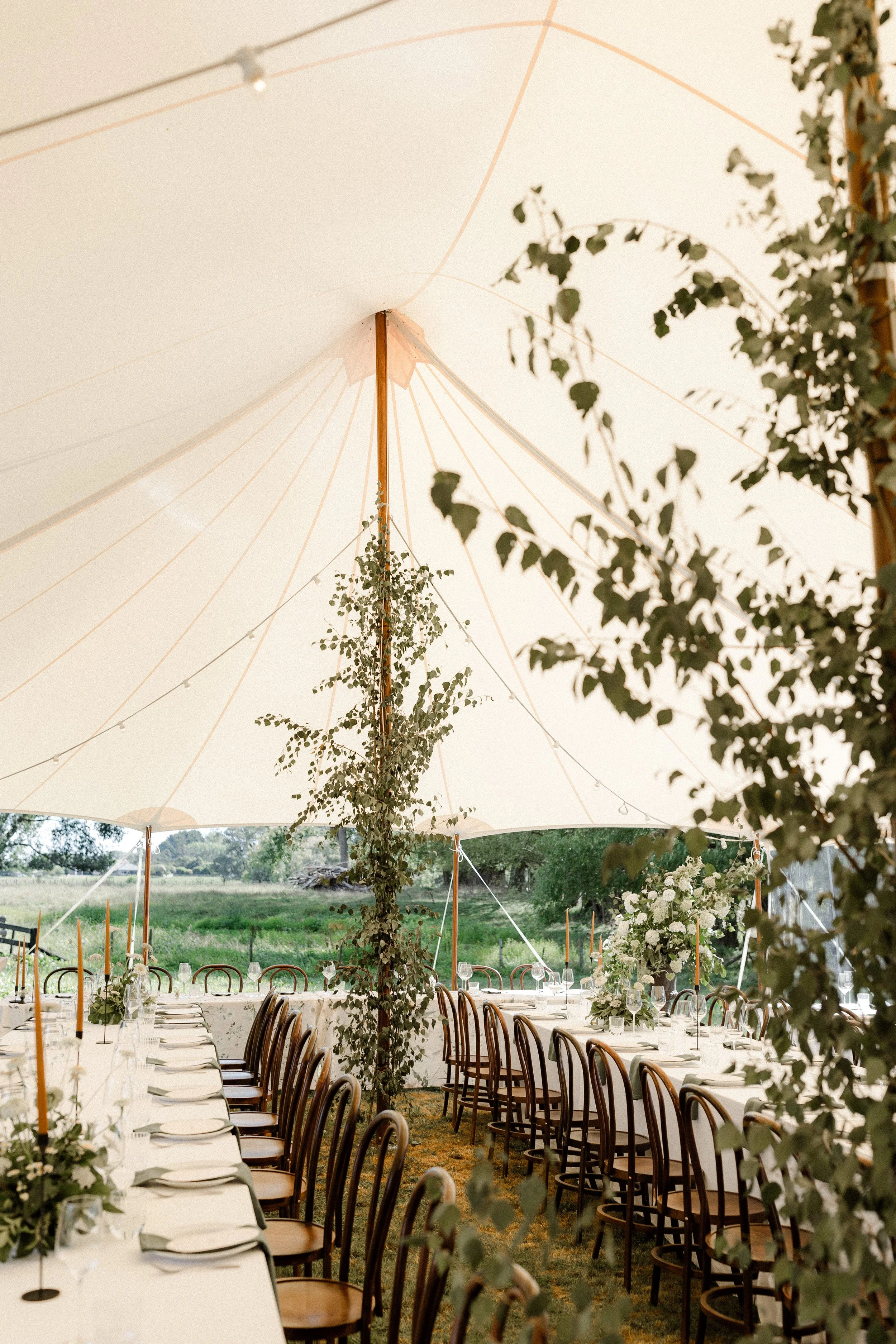 Outdoor wedding reception under a large white canopy with tables decorated with white tablecloths, candles, and floral arrangements, surrounded by greenery and trees.