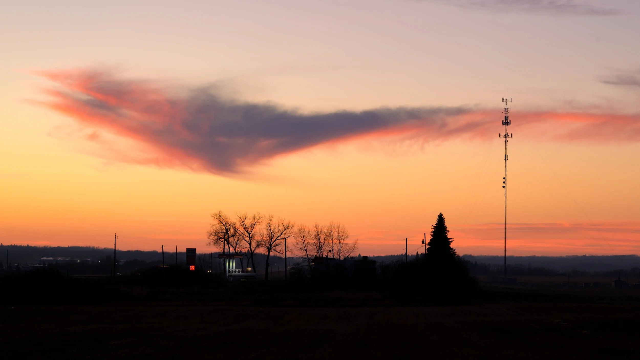 Sunset sky with orange and pink hues, featuring a large dark cloud, a tall radio tower, and silhouette trees in a rural landscape.
