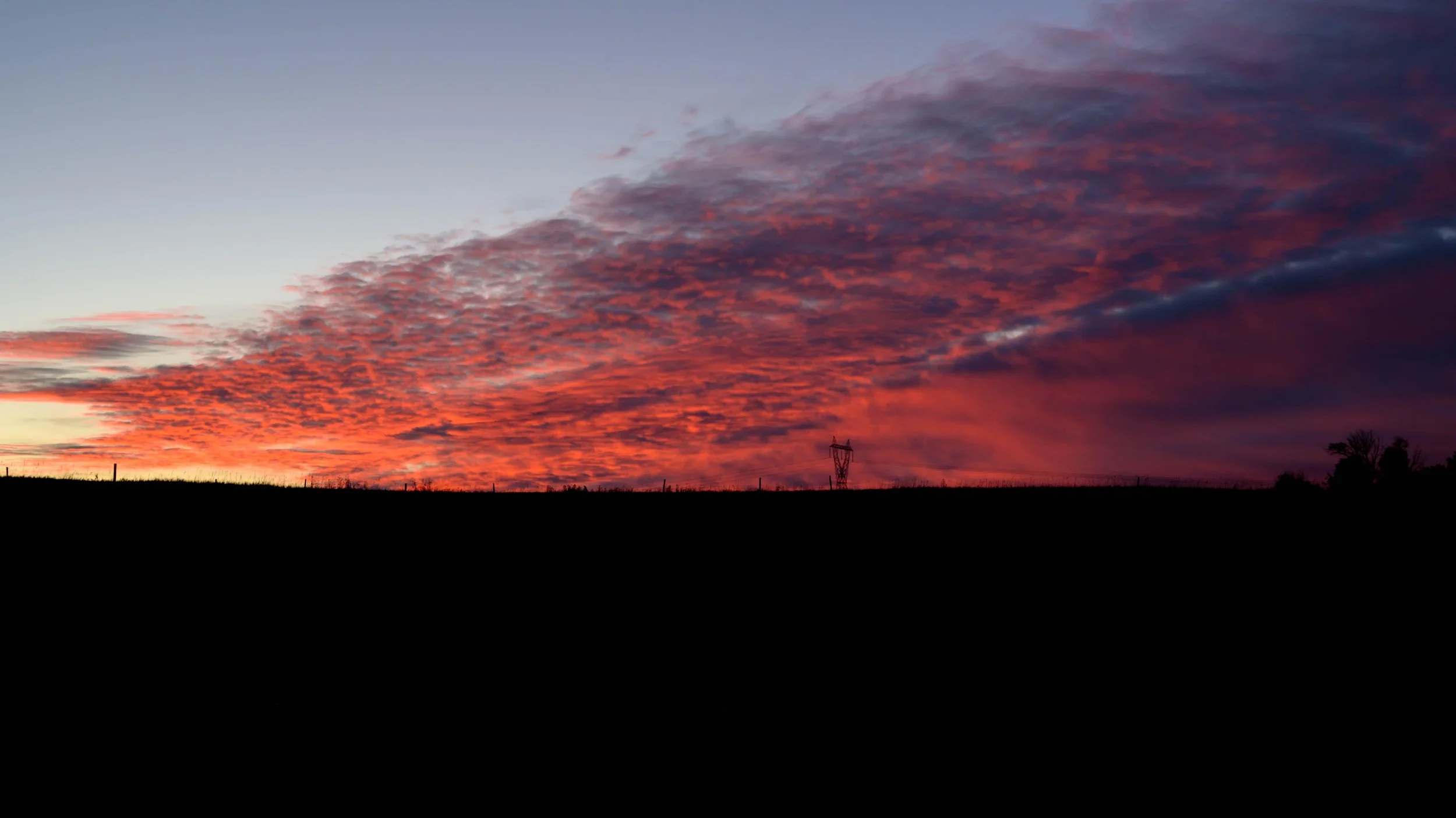 Colorful sunrise with orange, pink, purple, and dramatic clouds over a dark silhouette of a landscape with trees and power lines.