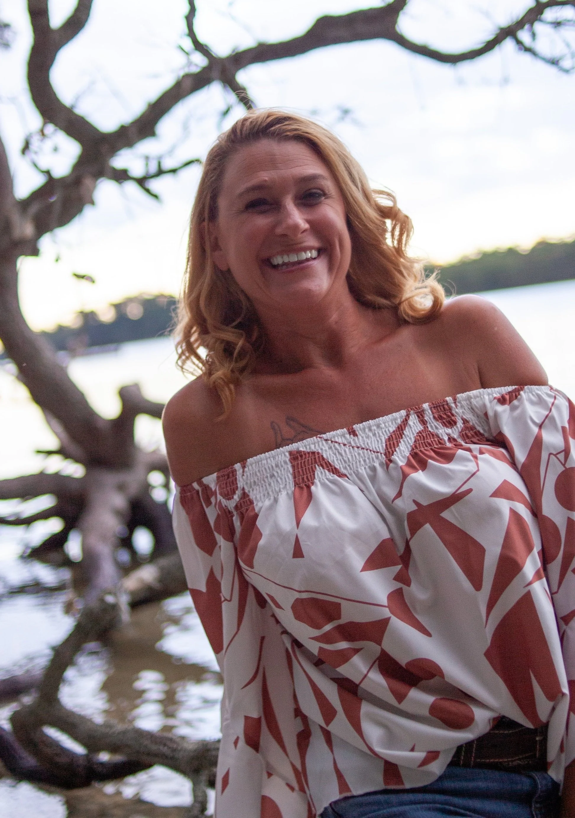 Smiling woman in patterned off-shoulder dress in outdoor setting with trees and water.