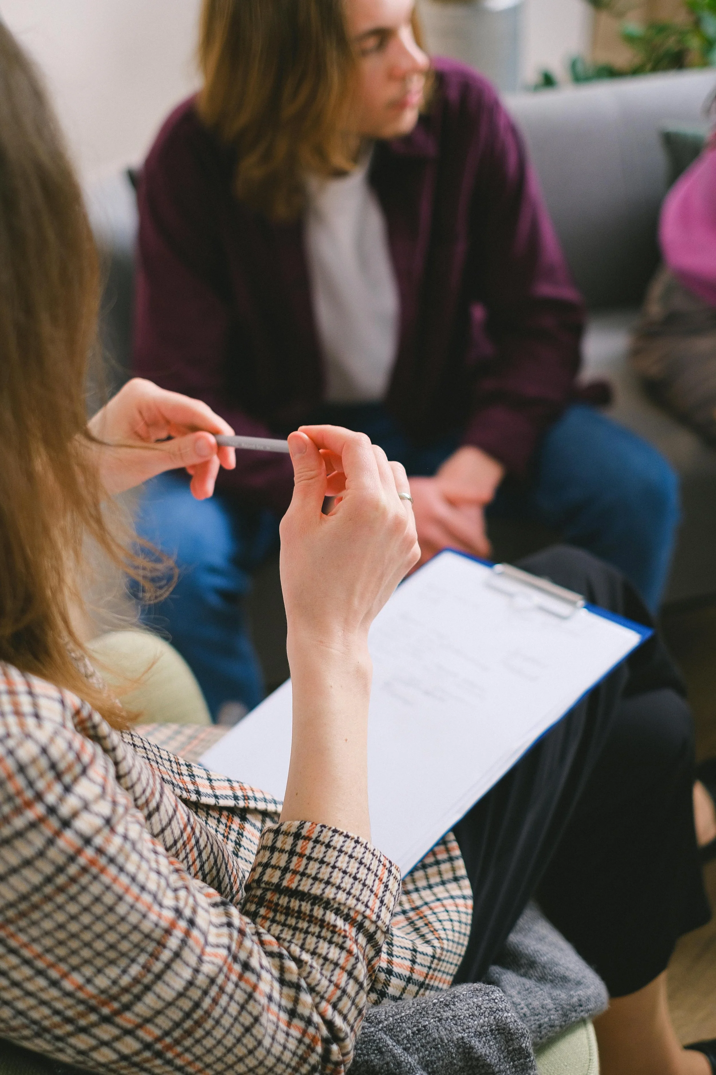 Person holding a pen and clipboard, sitting with another person in a casual setting.
