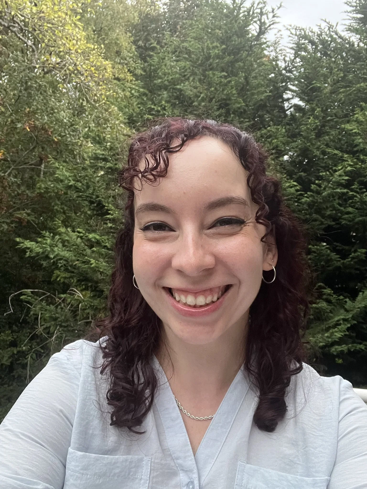A smiling woman with curly hair taking a selfie outdoors, surrounded by lush green trees.