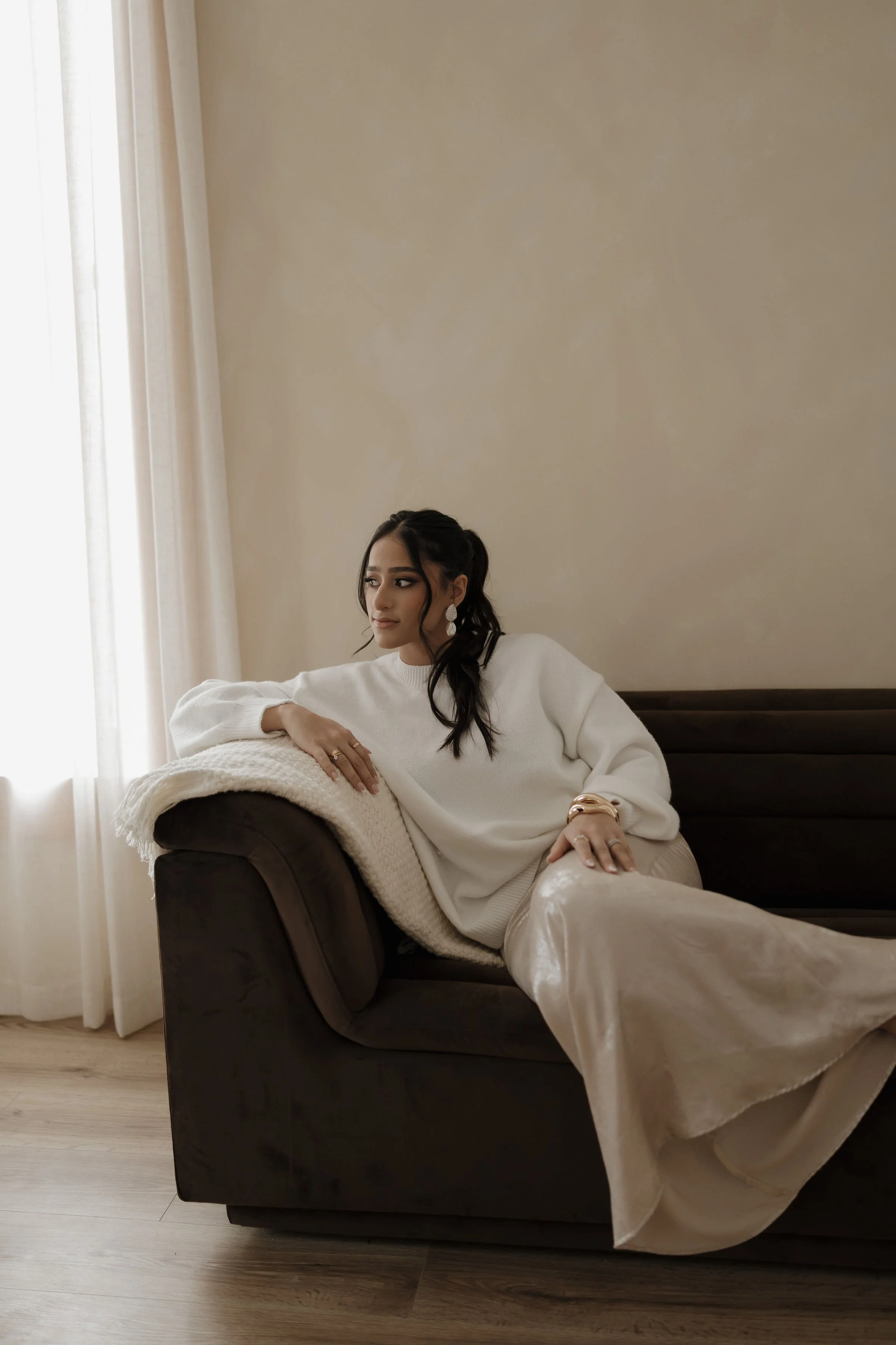 A woman with dark hair styled in loose waves, wearing a white oversized sweater and cream-colored satin pants, sits on a dark brown sofa in a room with beige walls and natural light coming through sheer curtains.