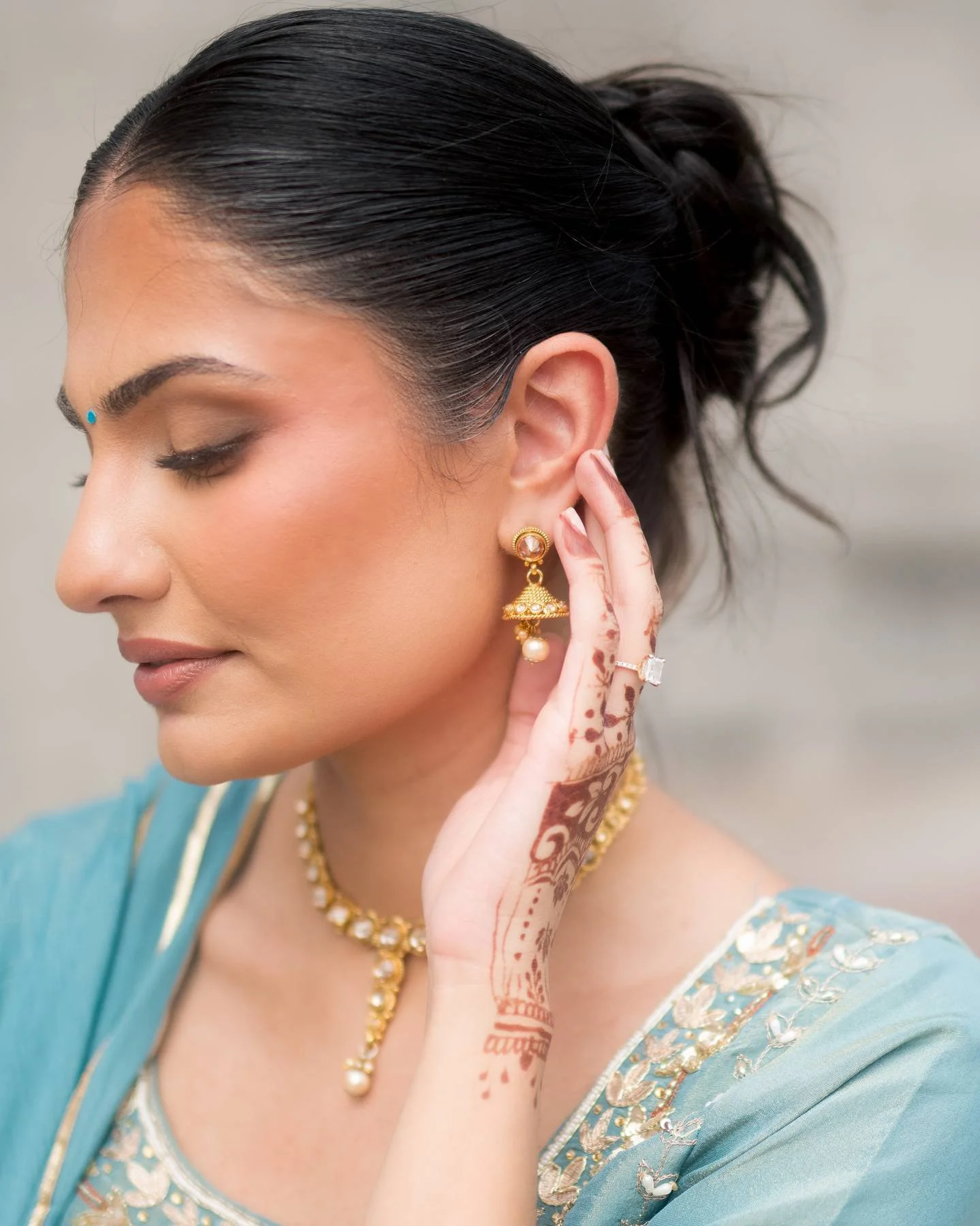 Close-up of a woman with dark hair styled in a bun, wearing traditional Indian jewelry, a blue sari with embroidery, and intricate henna designs on her hand, with her eyes closed.