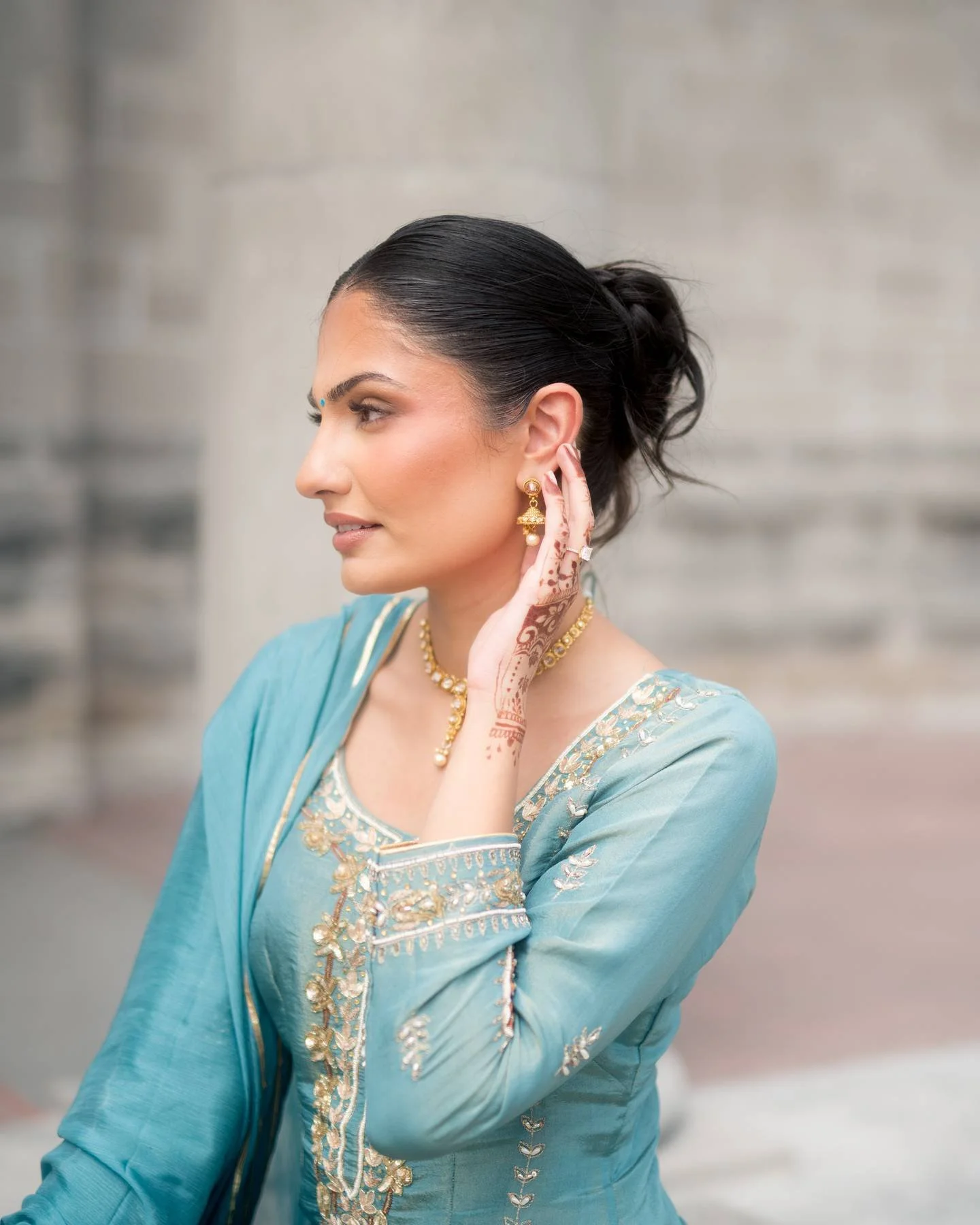 A woman dressed in a traditional blue and gold embroidered Indian outfit, with henna on her hand, wearing jewelry and earrings, touching her ear.