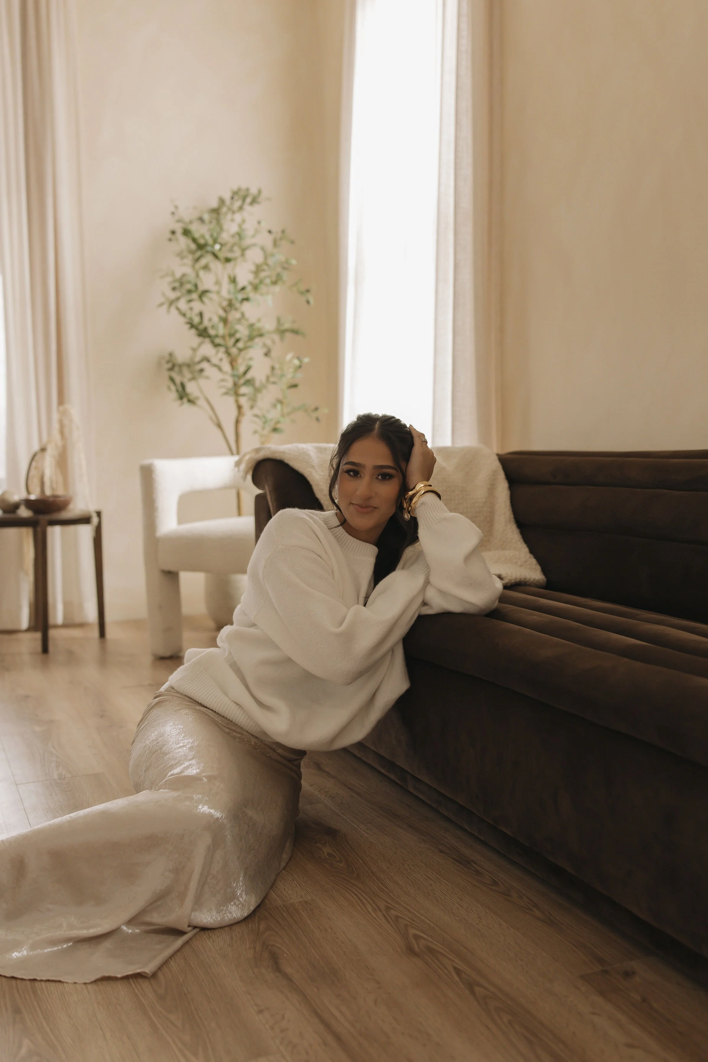 Young woman sitting on the floor of a living room, leaning against a dark brown sofa, with a large, beige, silky blanket on the floor. She is wearing a white sweater, beige pants, and gold jewelry, and is smiling softly at the camera. The room has light-colored walls, a cardboard and wood floor, and a potted plant near the window with cream curtains.