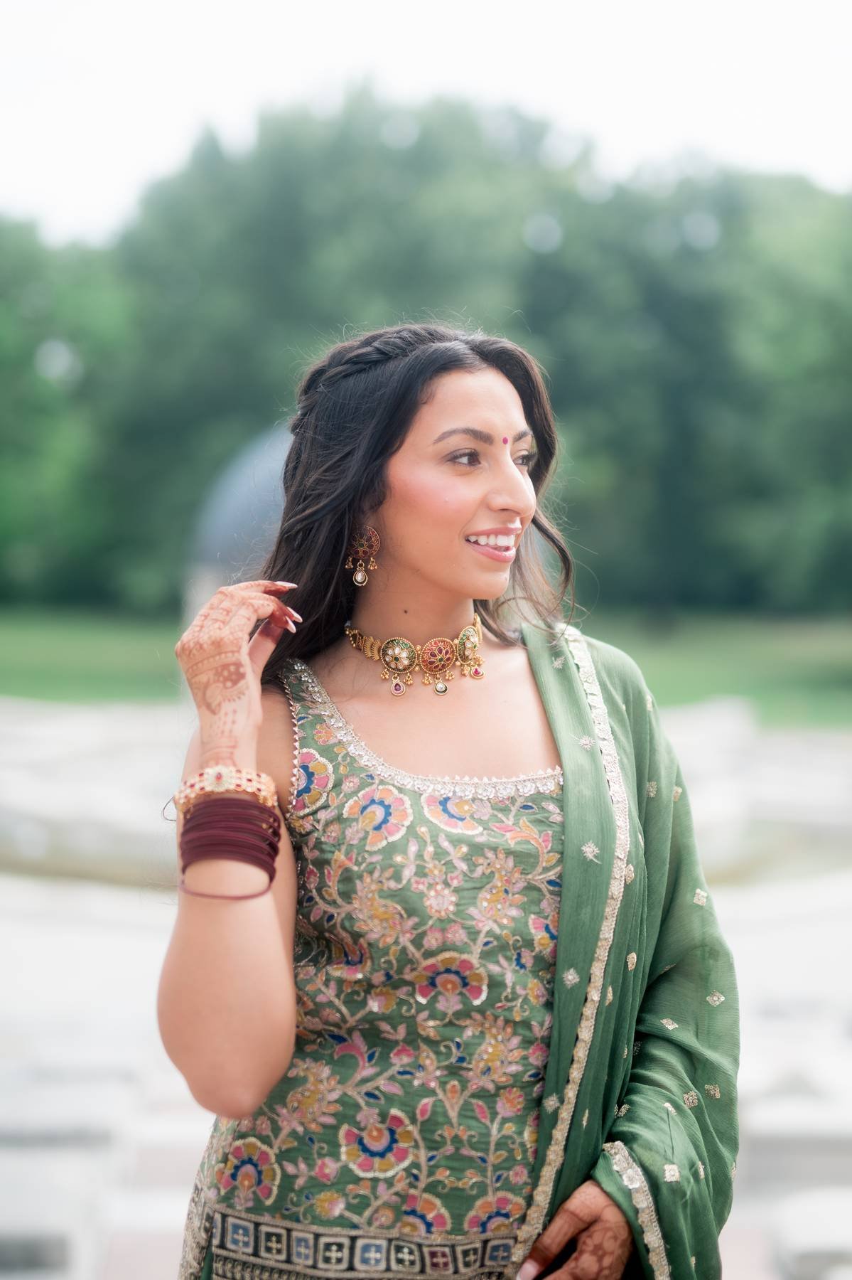 A woman dressed in a traditional Indian outfit, with intricate jewelry and henna on her hand, standing outdoors near water with green trees in the background.
