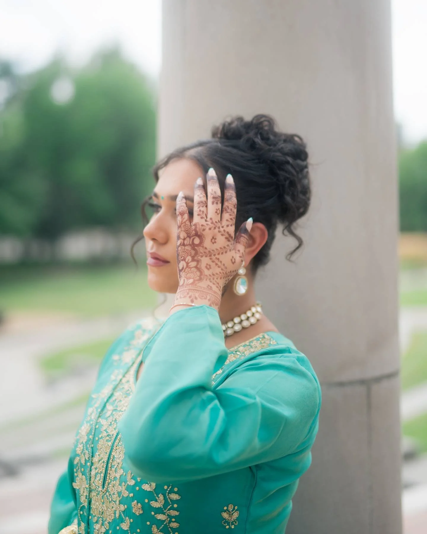 A woman dressed in traditional Indian attire with henna designs on her hand, touching her forehead, standing outdoors with trees in the background.