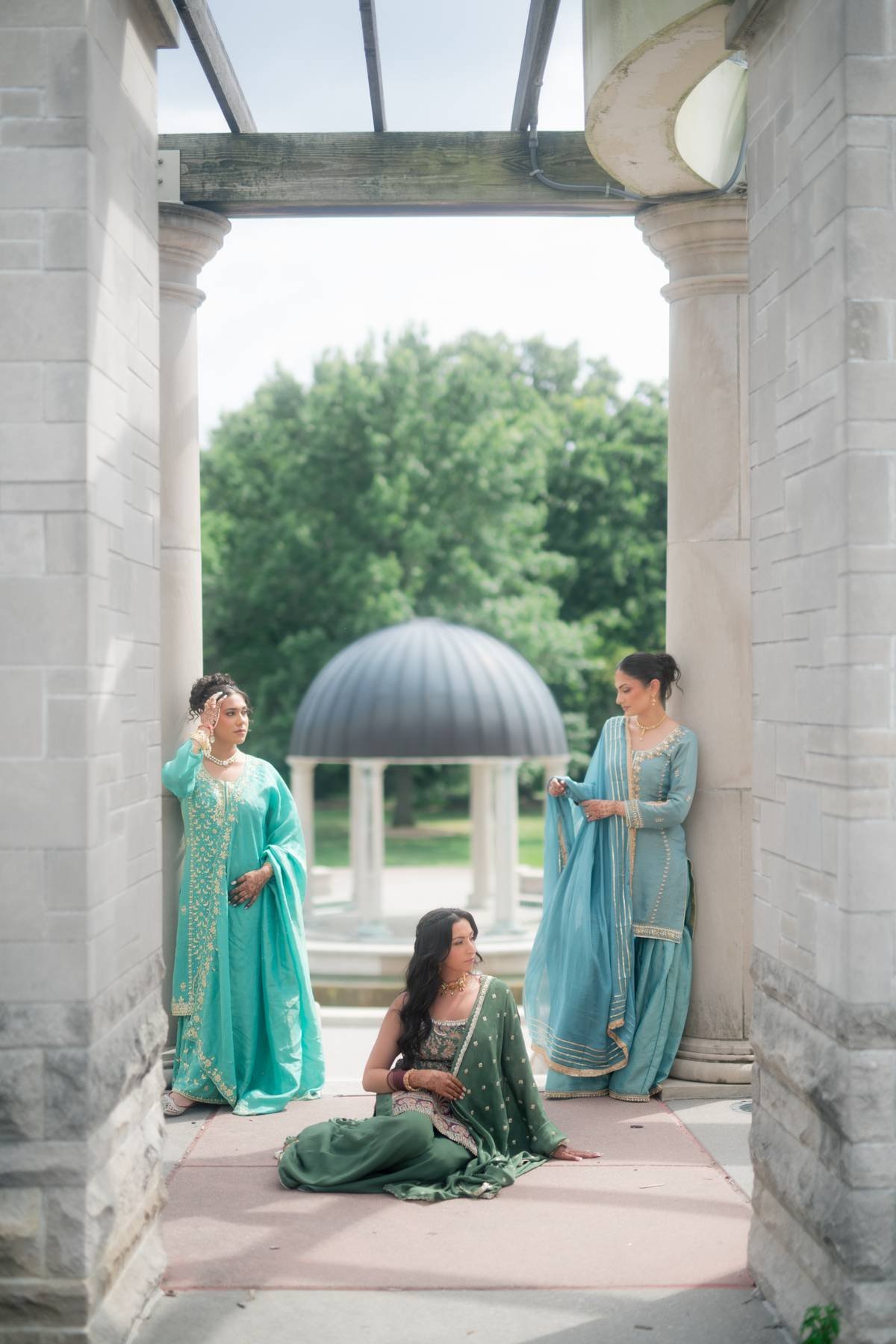 Four women in traditional South Asian clothing pose and relax in a classical architectural setting with stone columns, greenery, and a domed pavilion in the background.