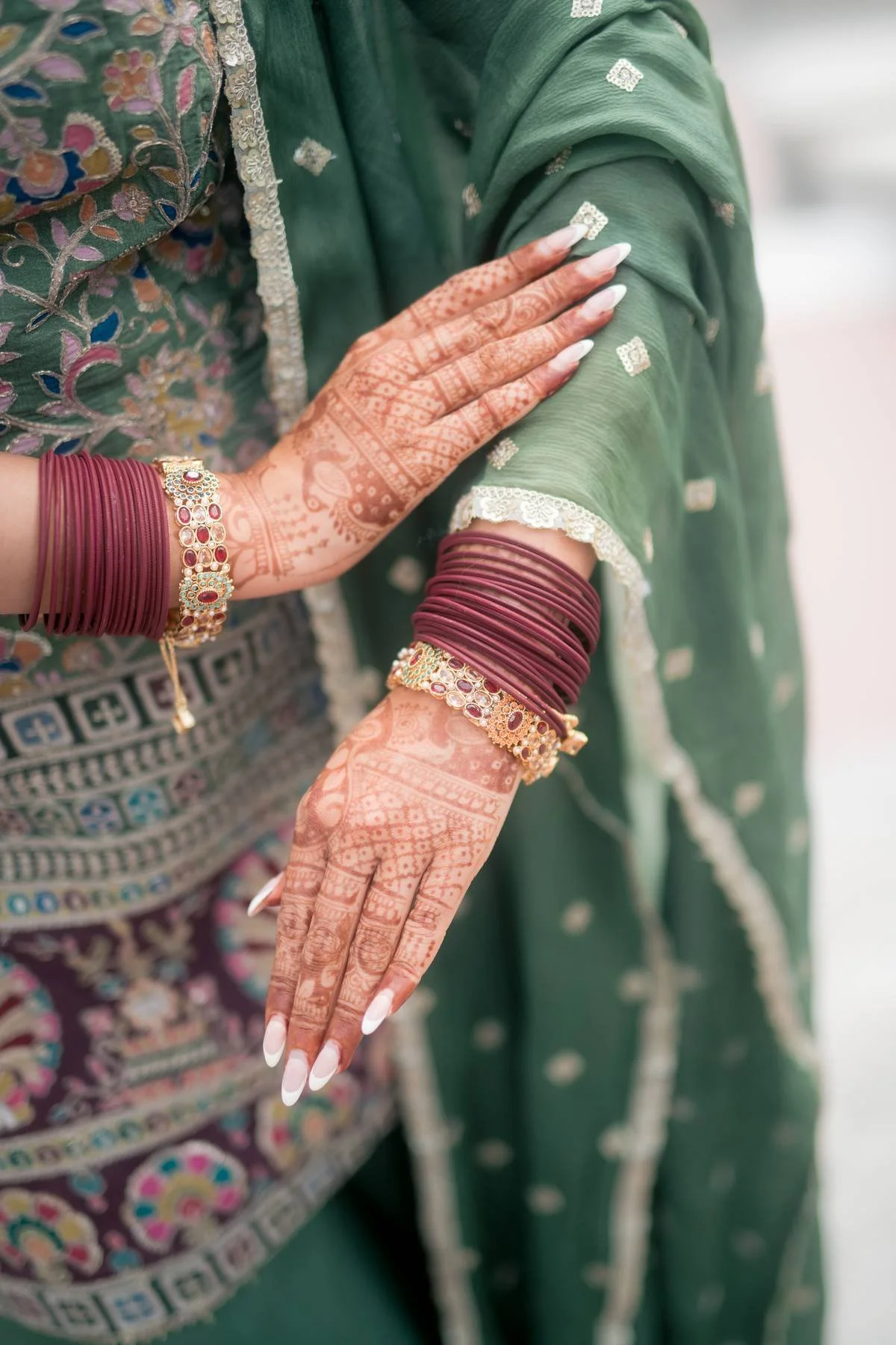 A woman wearing traditional Indian attire with intricate henna designs on her hands, adorned with red bangles and gold jewelry, is adjusting or holding the sleeve of her green embroidered dress.