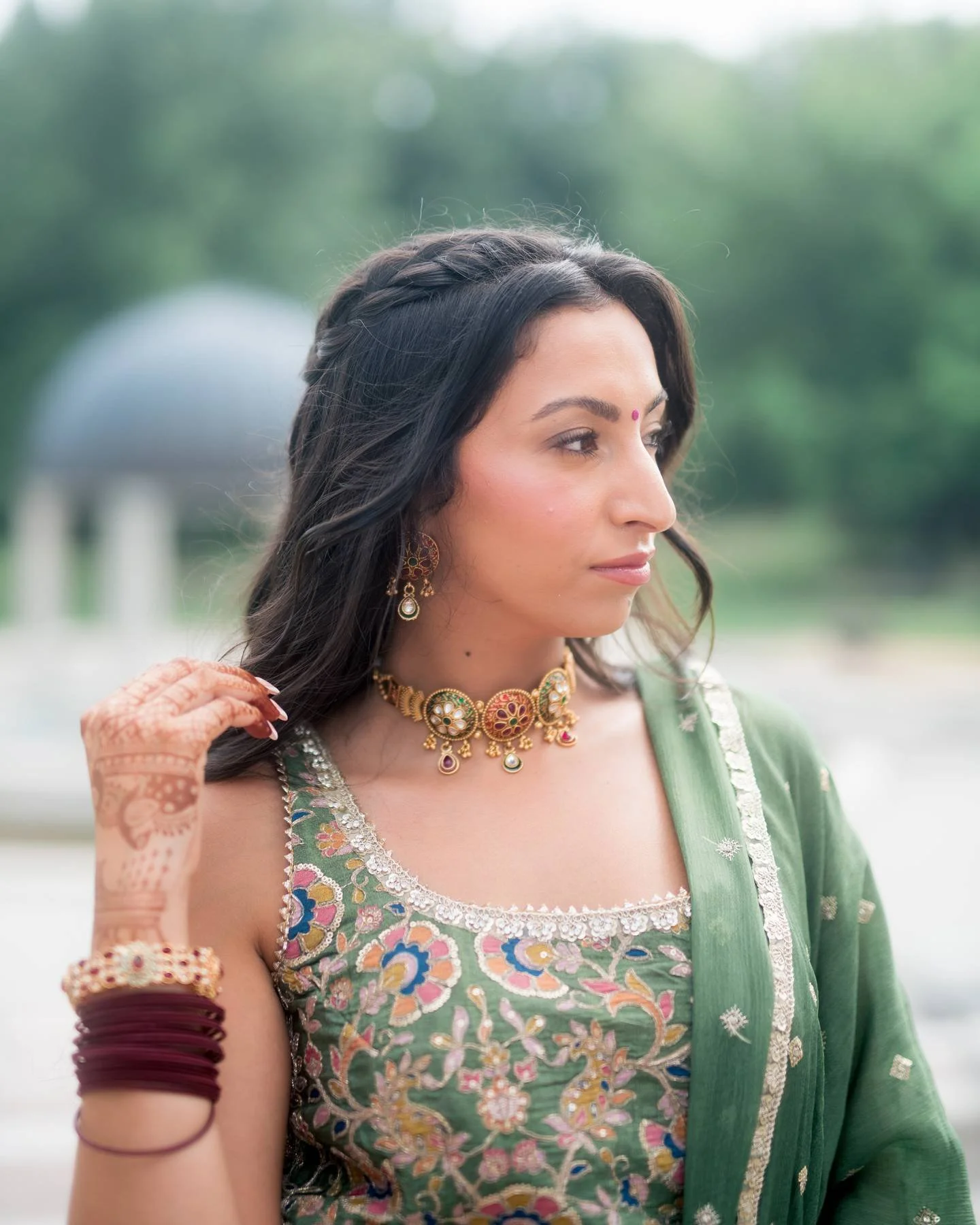A woman wearing traditional Indian attire and jewelry, with henna on her hand, outdoors with greenery in the background.
