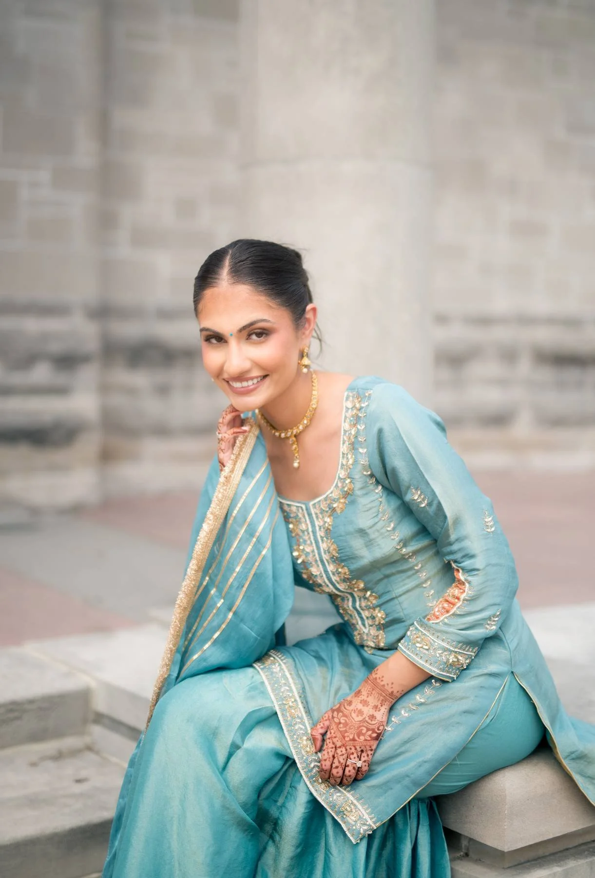 Woman dressed in blue traditional Indian attire, sitting on steps, smiling, with jewelry, henna on hand, and a stone wall background.