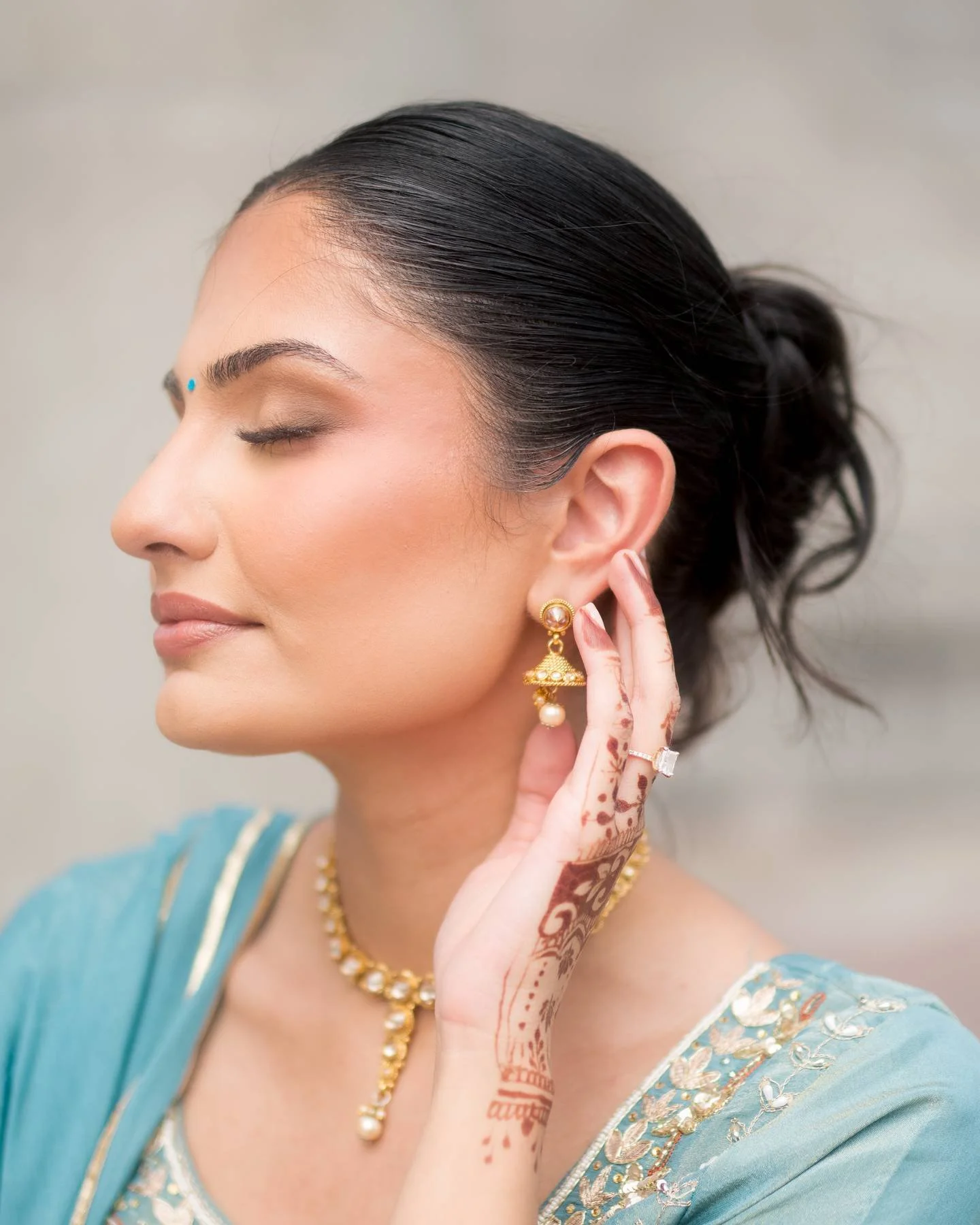 A woman with closed eyes, wearing traditional Indian jewelry and a blue saree, with henna designs on her hand and face, looks serene.